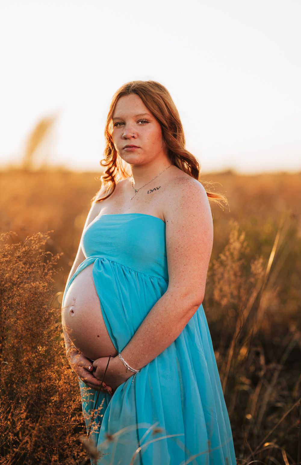 Pregnant woman in turquoise dress poses in golden sunlit field during sunset maternity photoshoot.