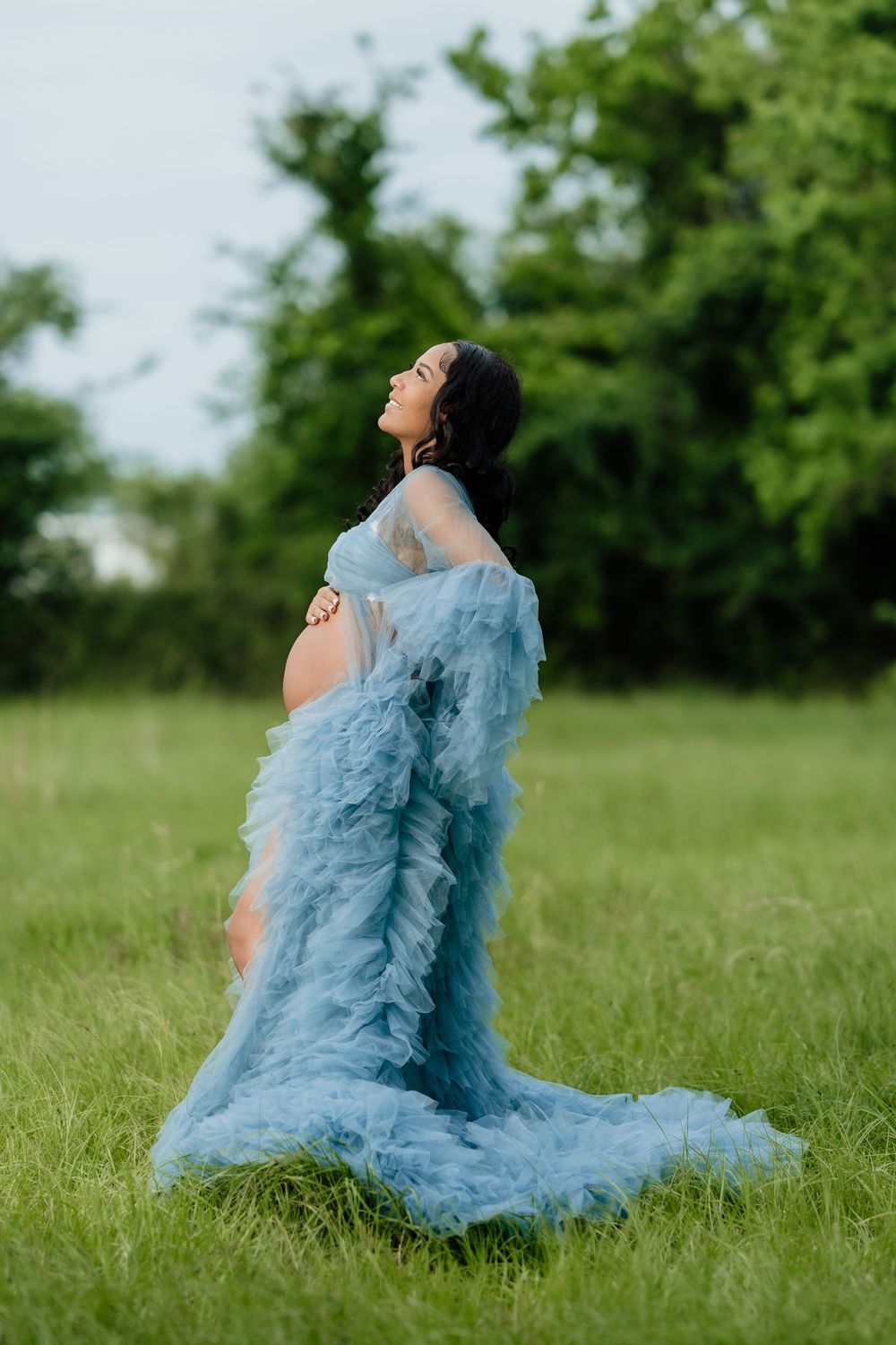 Pregnant woman in flowing blue tulle gown standing in green field, cradling baby bump during outdoor maternity photoshoot.