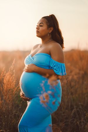 Expectant mother in flowing blue off-shoulder dress poses serenely in golden field at sunset.