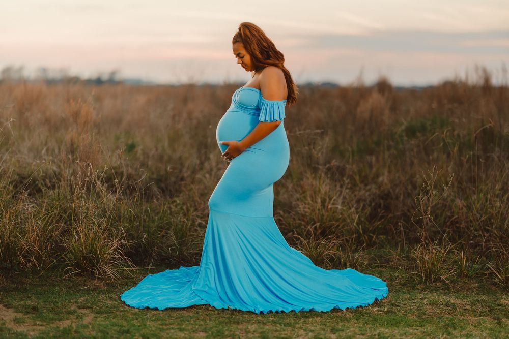 A pregnant woman in a flowing turquoise gown poses in a field at sunset, cradling her belly.