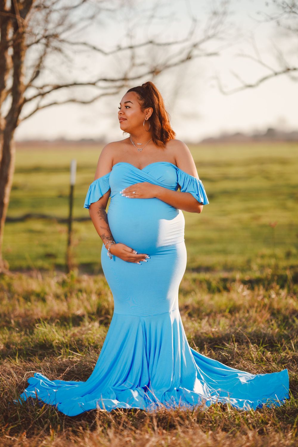 A pregnant woman in a flowing light blue off-shoulder gown poses gracefully in a sunlit field at golden hour.