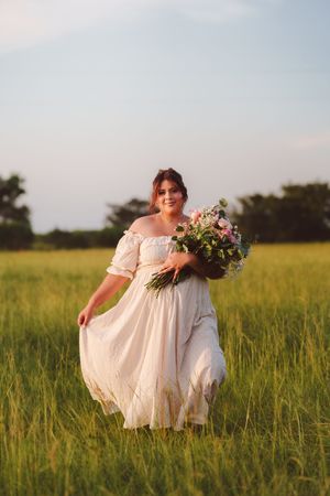 A bride in an off-shoulder white dress carries a floral bouquet while standing in a sunlit grassy meadow at sunset.