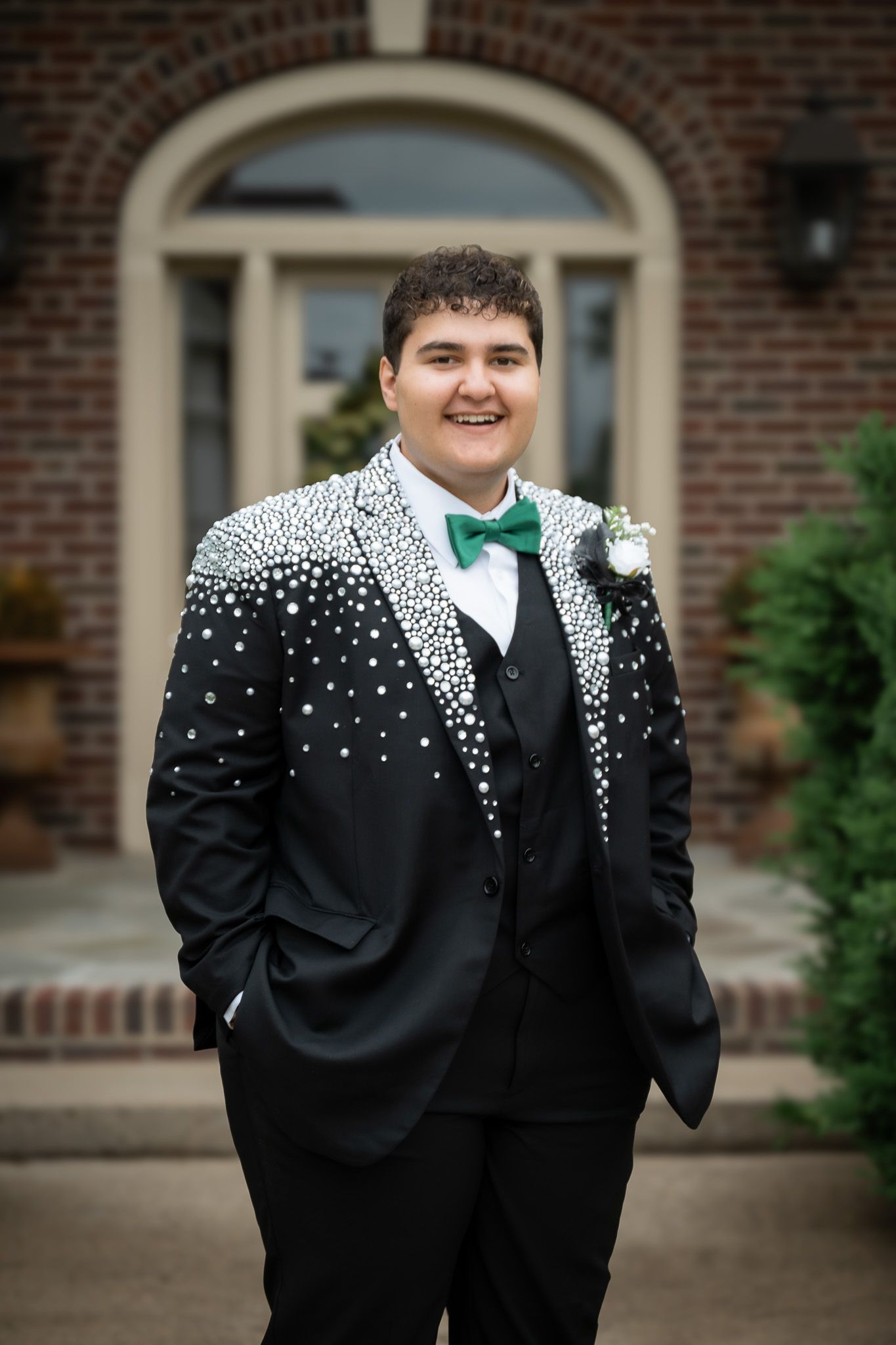 A formal black tuxedo with a green bowtie and unique white scattered dot pattern stands before a brick entrance.