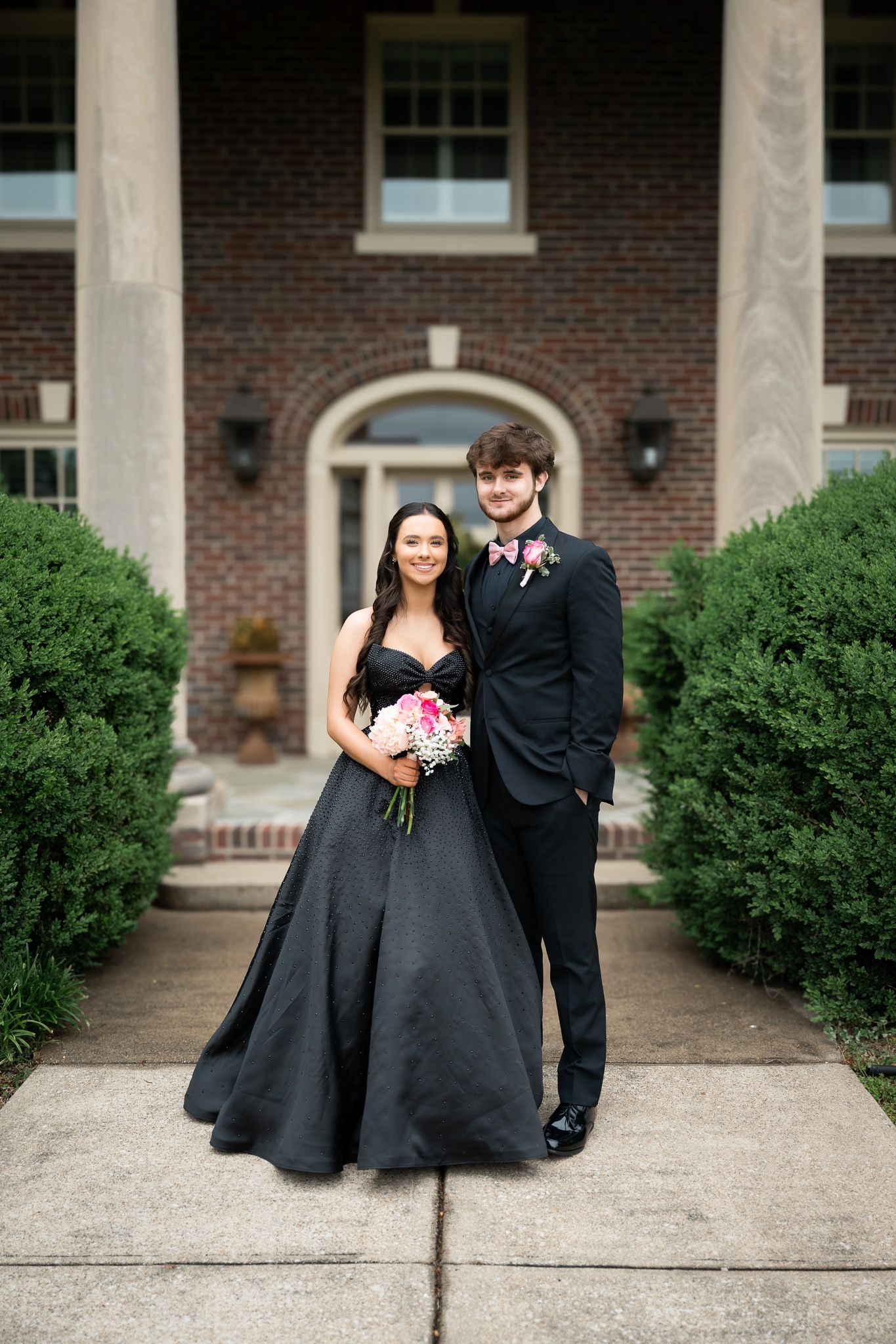 Two people in formal attire pose together for a photo outside a brick building with white pillars.