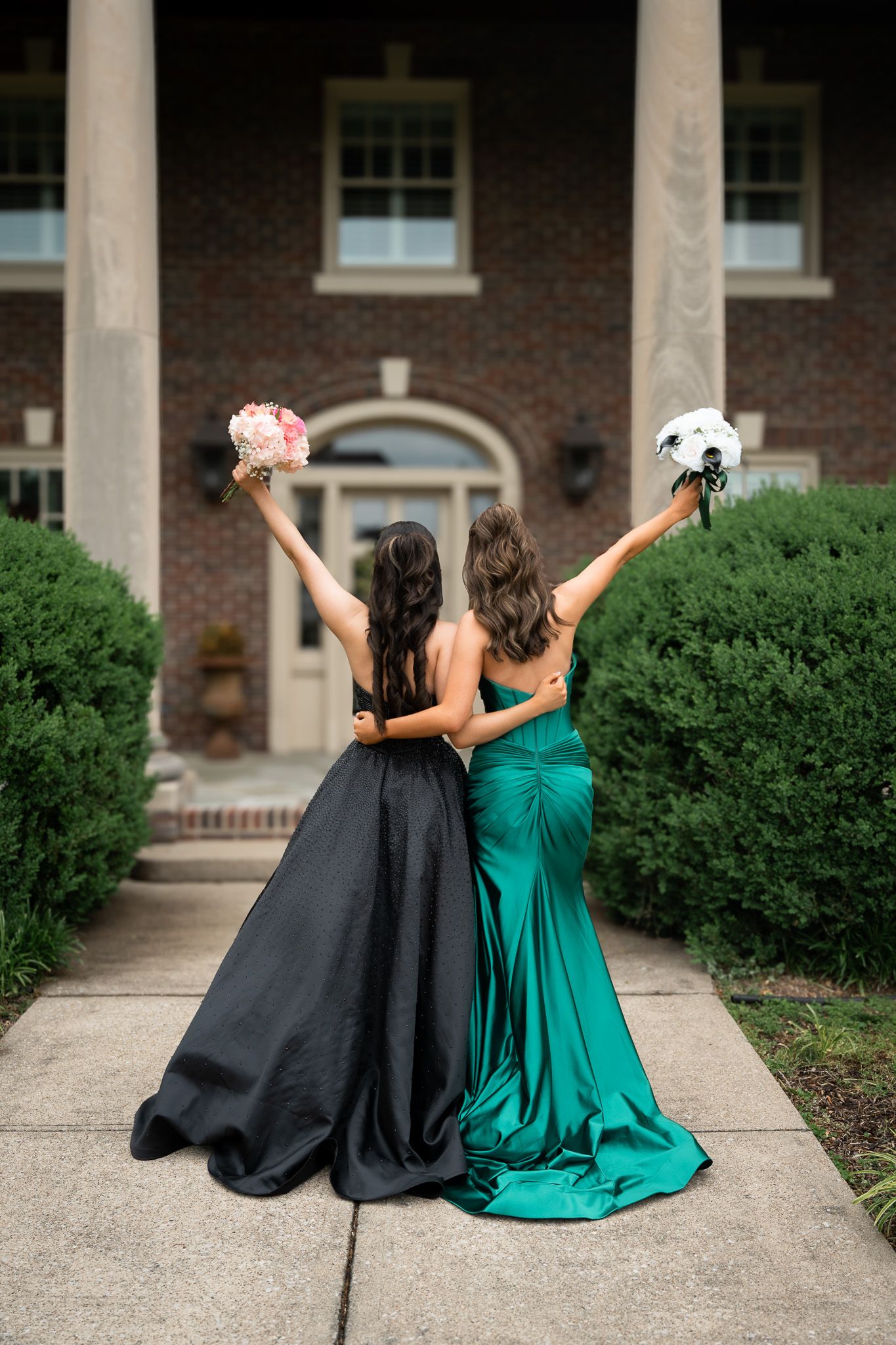 Two women in formal dresses, one black and one emerald green, raise their bouquets triumphantly outside a brick building.