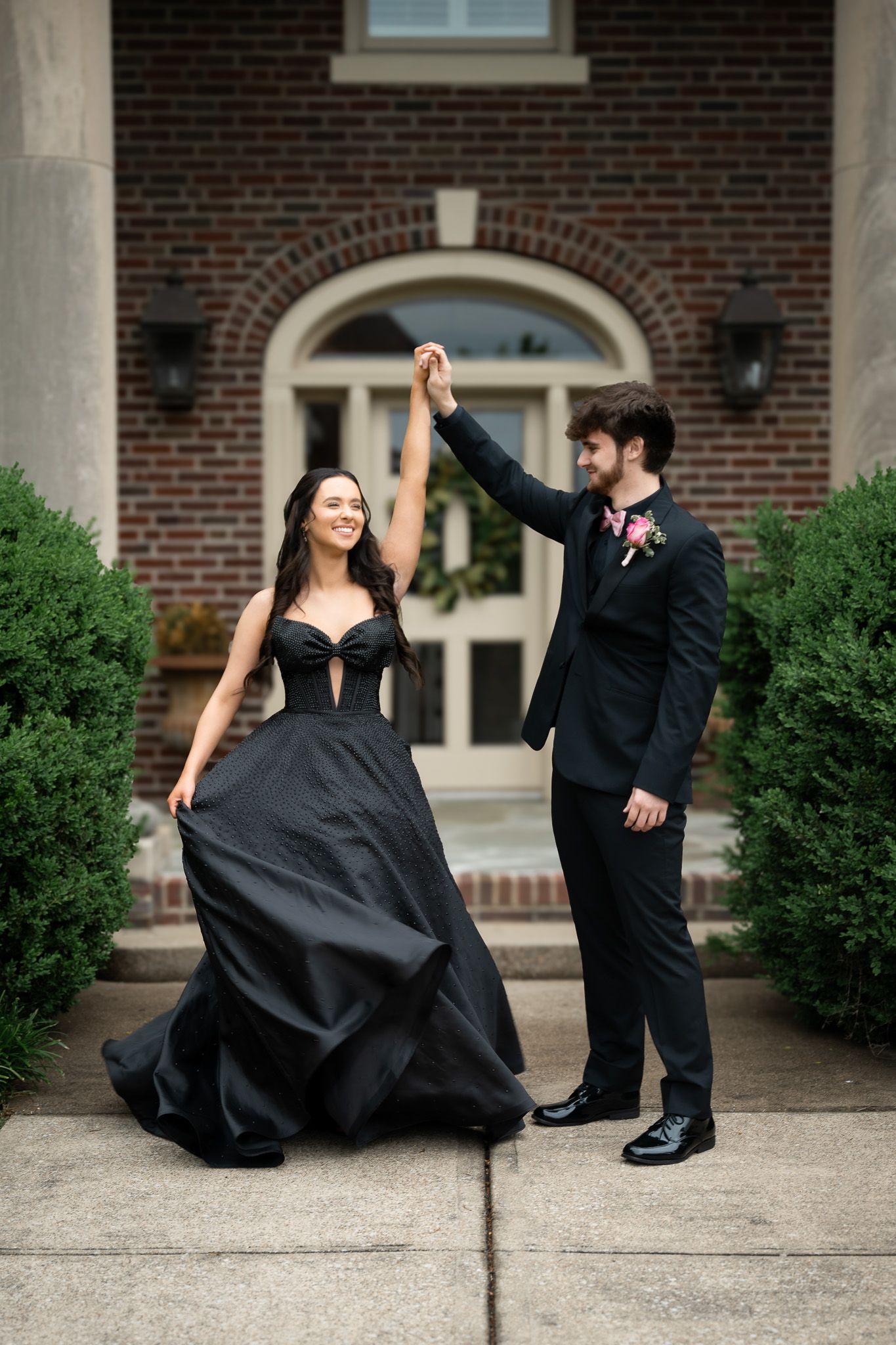 A couple in formal attire dancing together on a brick building entrance walkway lined with greenery.