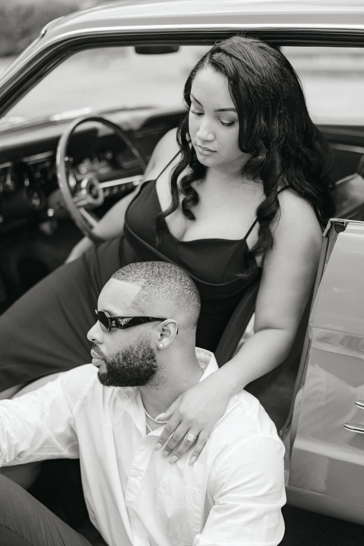 Black and white photo of a couple posing with a vintage car, woman in black dress resting hand on mans shoulder.