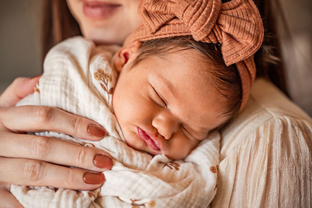 Sleeping newborn in a brown headband rests peacefully against caregiver wearing white.