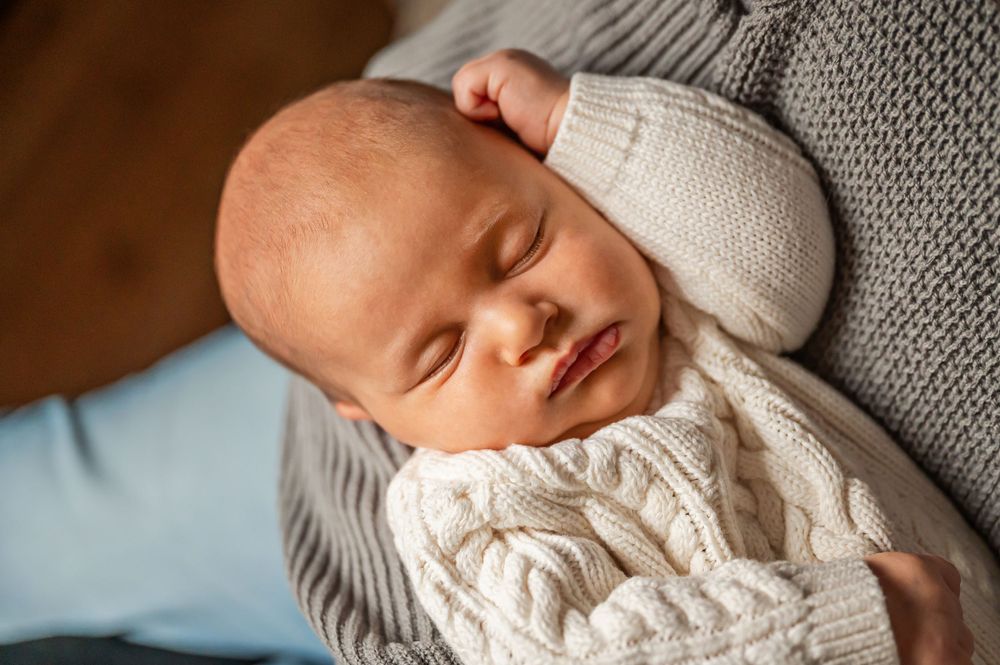 A sleeping newborn in a white cable knit sweater rests peacefully against grey knit fabric.