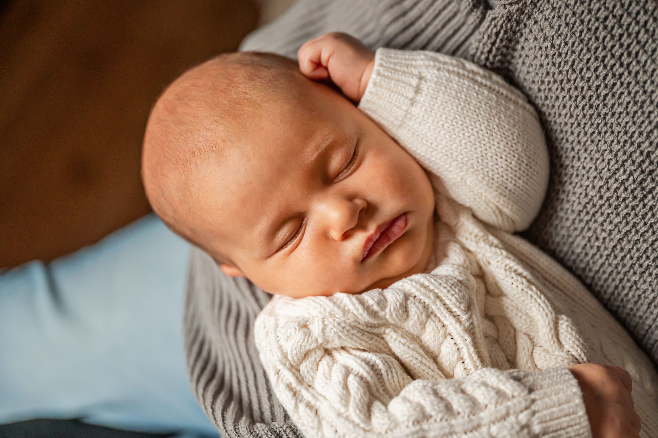 A sleeping newborn in a white cable knit sweater rests peacefully against grey knit fabric.