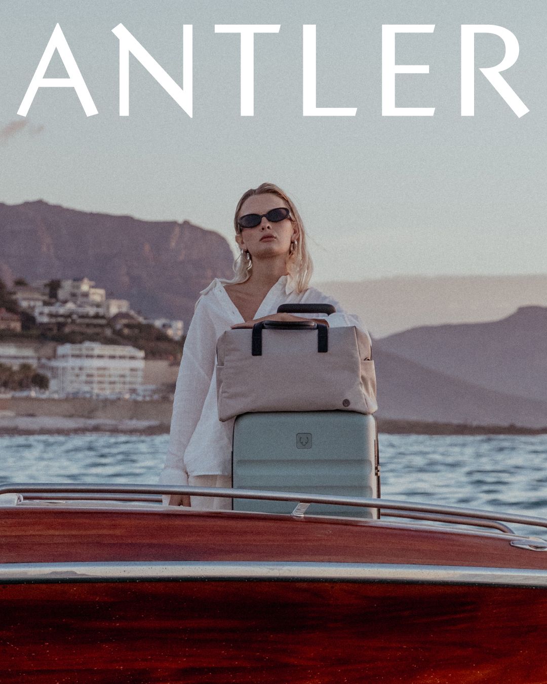 Woman standing on vintage wooden boat carrying Antler luggage bag at sunset with mountains in background.