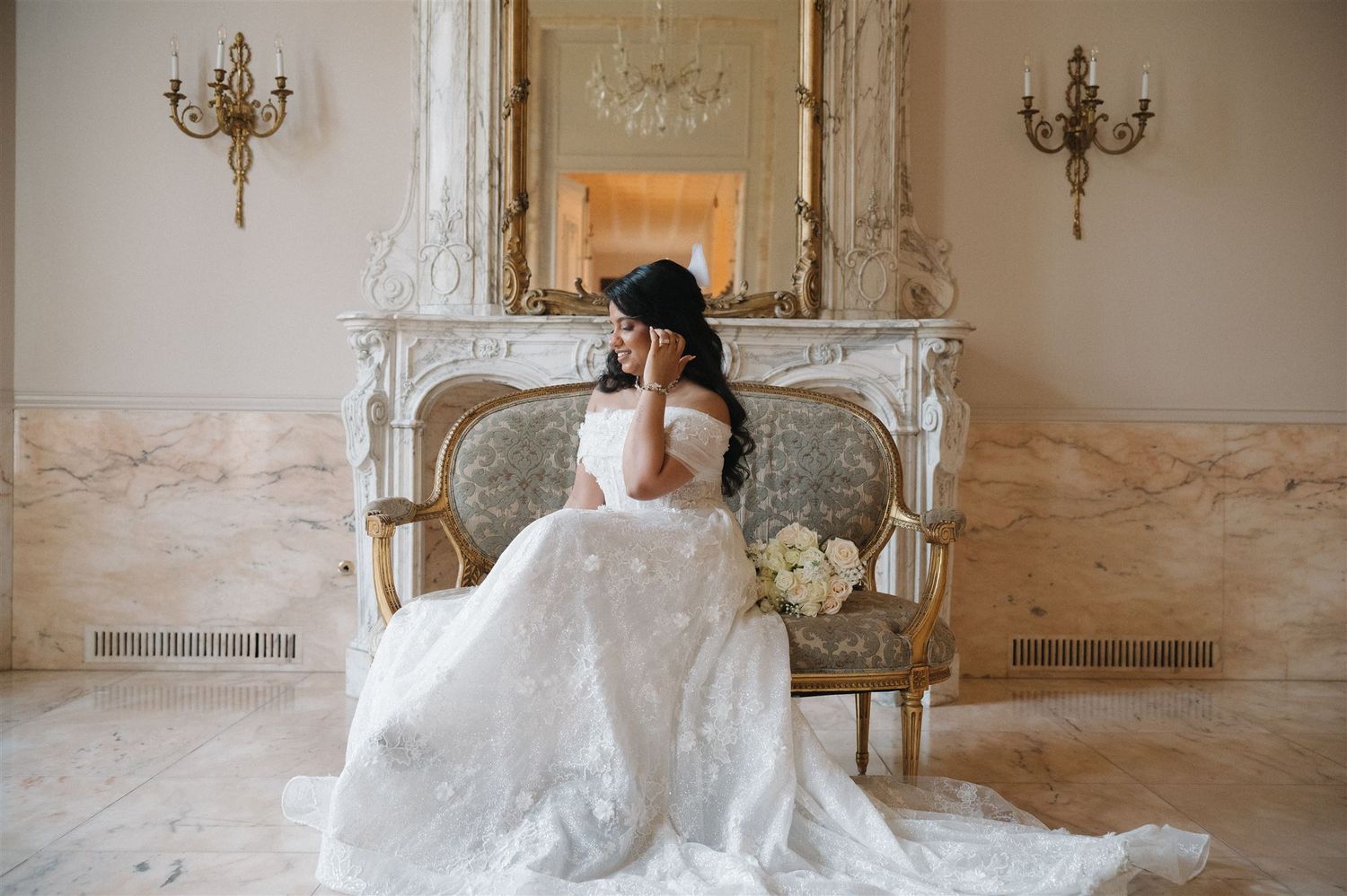 A bride wearing an elegant white lace wedding gown on antique sofa in luxurious marble room with ornate details.