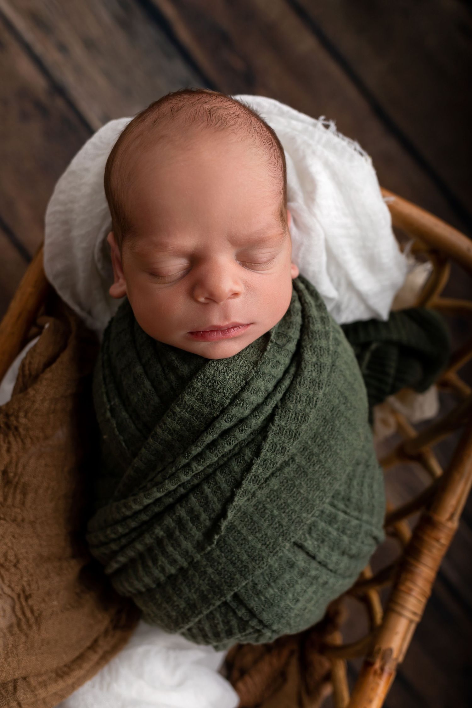 Sleeping newborn wrapped in green swaddle blanket resting in wooden basket.