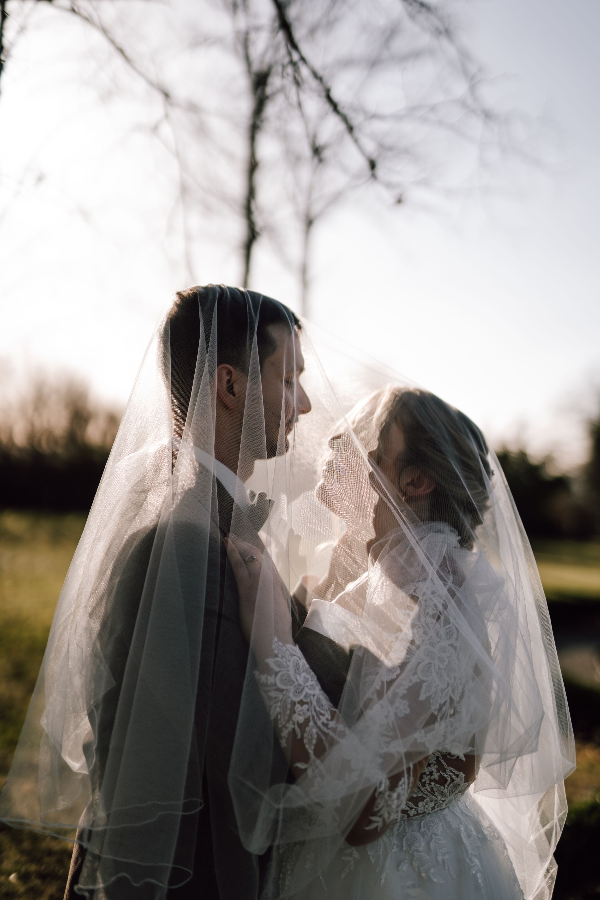 Romantic wedding couple embracing under a flowing white veil against a natural outdoor backdrop.