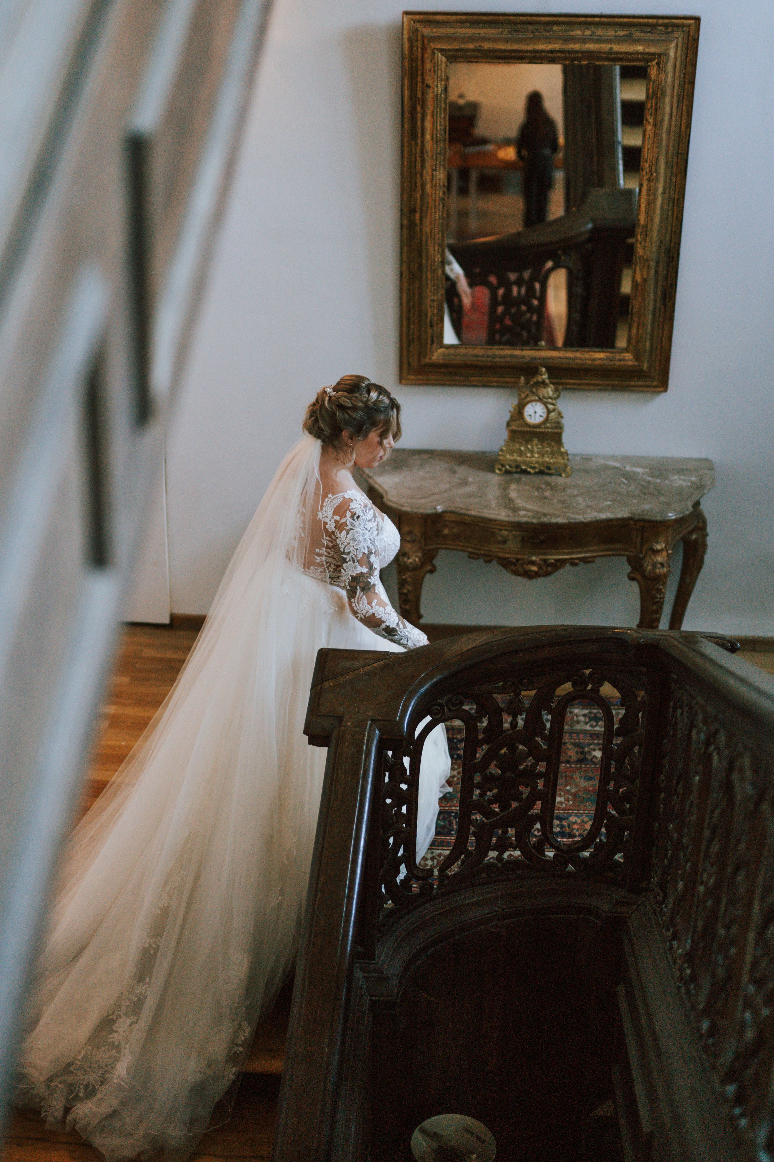 Elegant bridal gown flowing down ornate staircase with vintage mirror and decor.