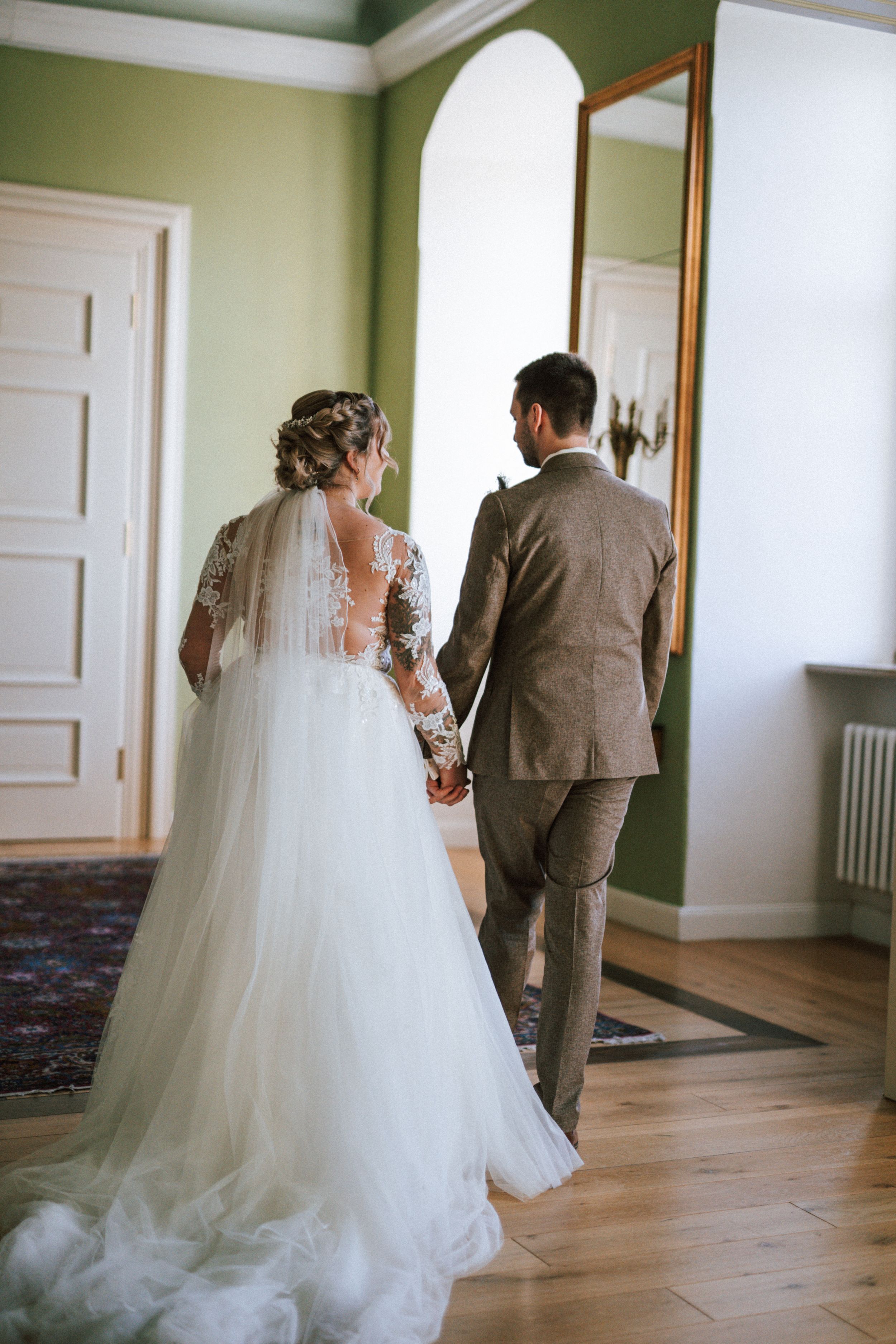 A bride and groom walk through an elegant green-walled room with hardwood floors and white trim.
