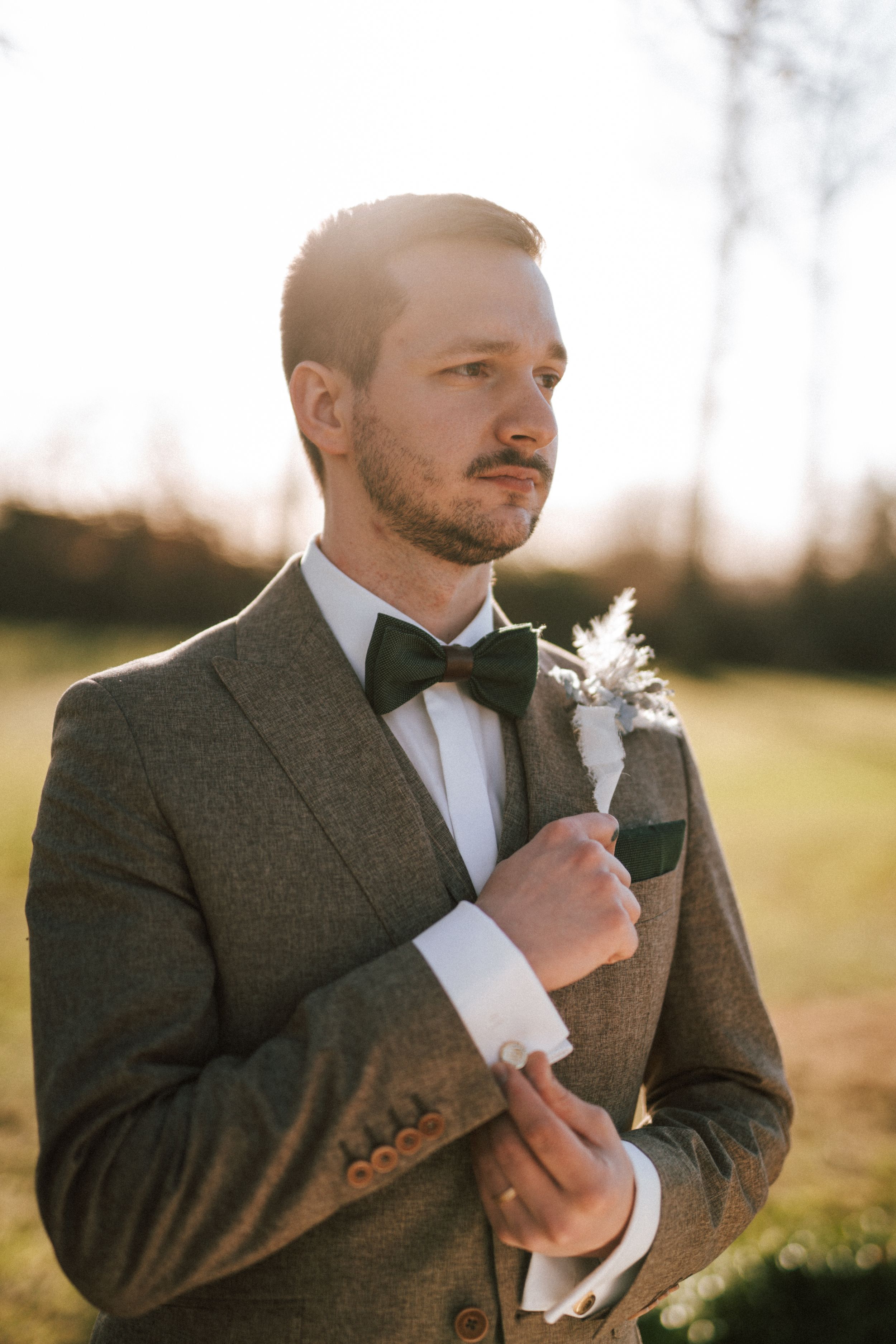 Groom in tweed suit adjusts his bowtie during golden hour.