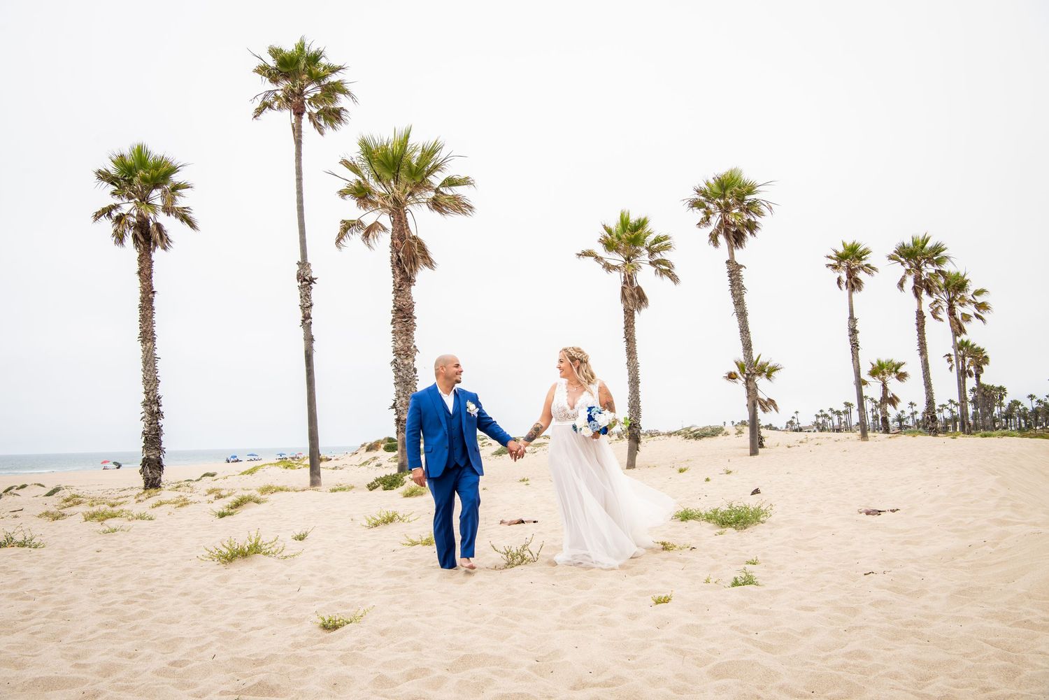 Newlyweds walk joyfully on beach lined with tall palm trees.