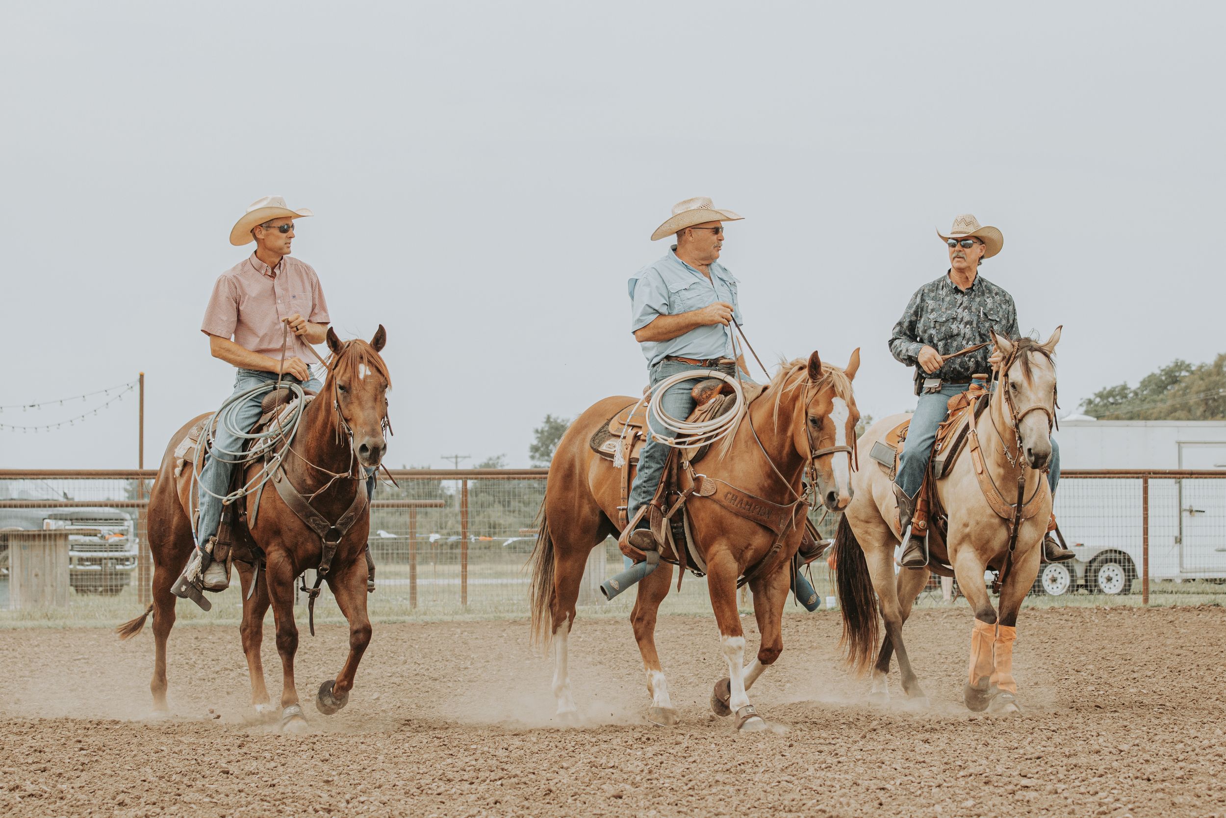 Three riders on horseback gallop side by side in dusty arena.