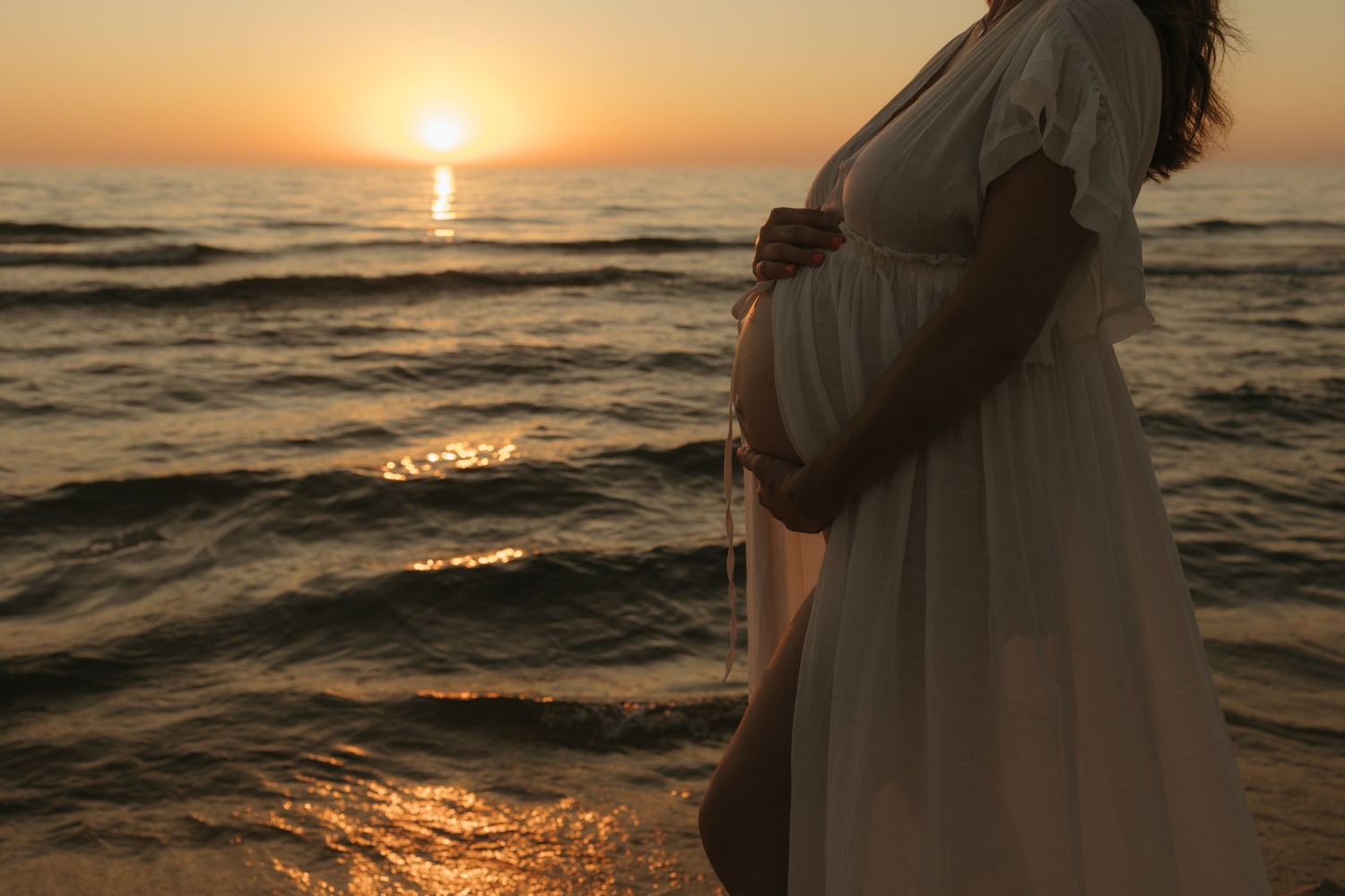 Silhouette of pregnant woman in white dress standing by ocean waves during golden hour for maternity photos.