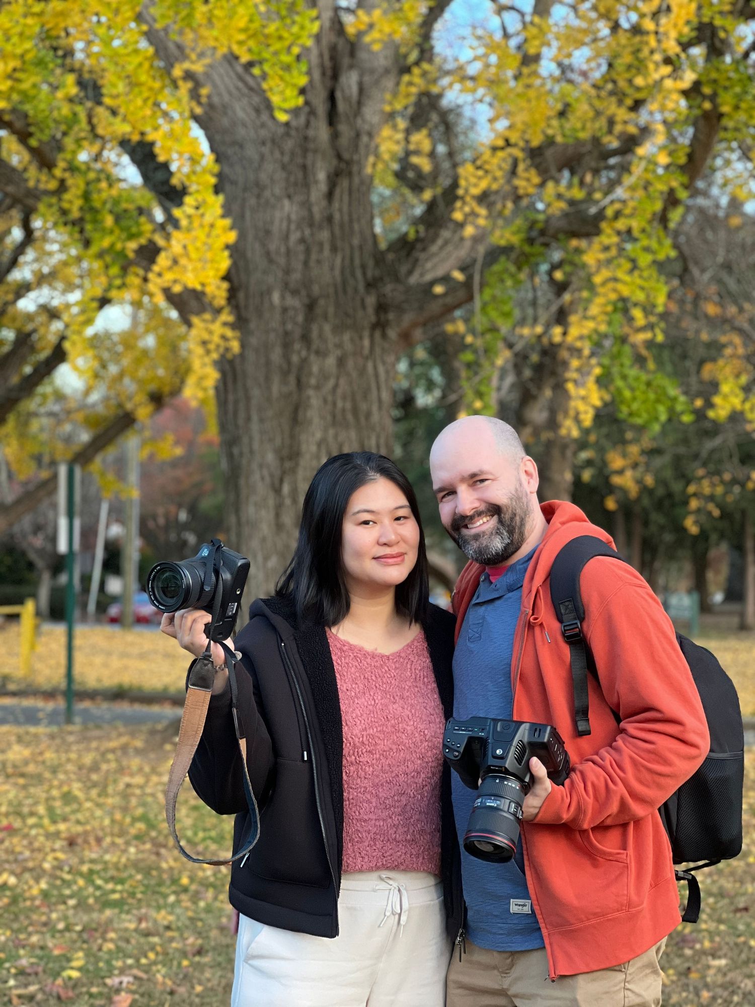 Two photographers with professional cameras stand together among fall foliage in a park.