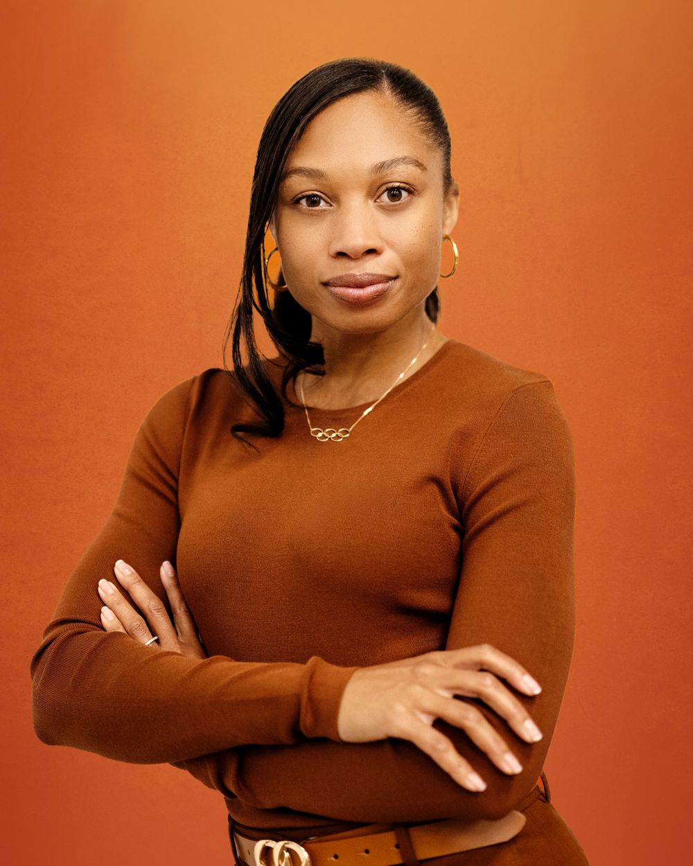 Allyson Felix. Professional portrait against an orange background showing person in brown sweater with gold jewelry and styled hair.