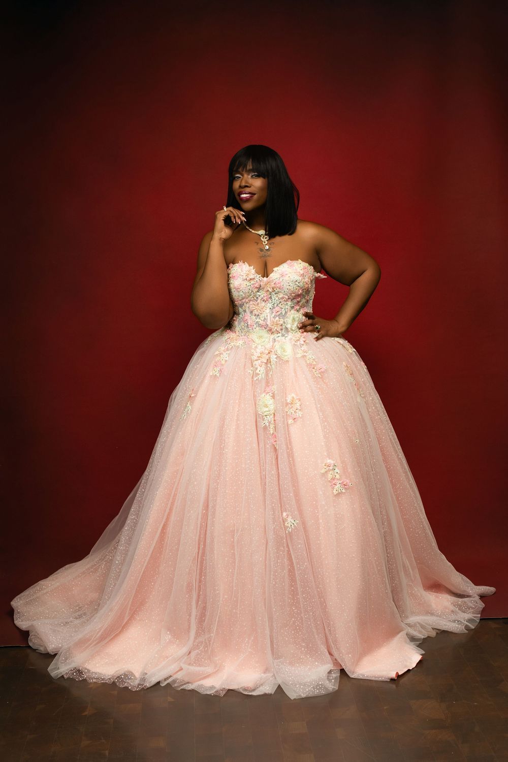 A person poses gracefully in a flowing pink tulle ball gown against a dark red backdrop.