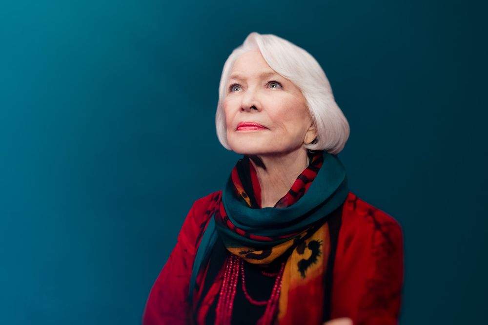 A woman with white hair wearing a red coat and teal scarf gazes upward against a teal background.