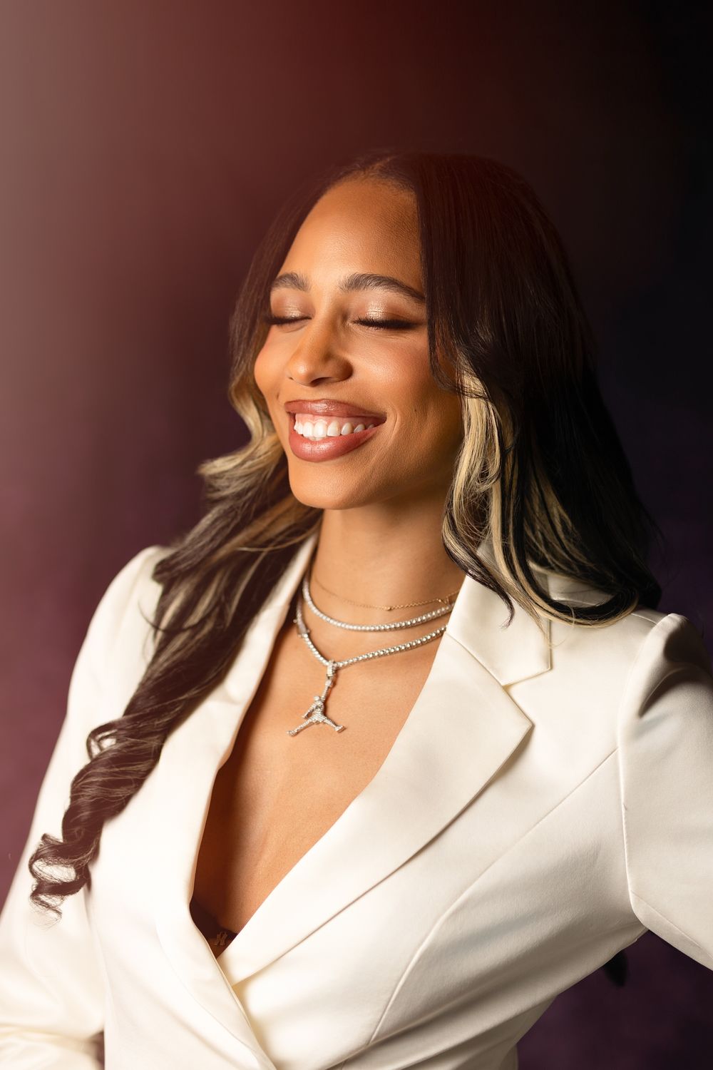 Smiling woman in white blazer with diamond necklace, eyes closed, long dark hair with highlights, against dark background.
