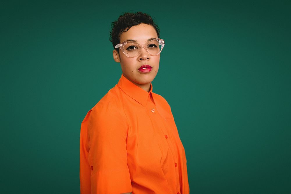 Person in orange shirt and clear glasses poses against a dark green background.