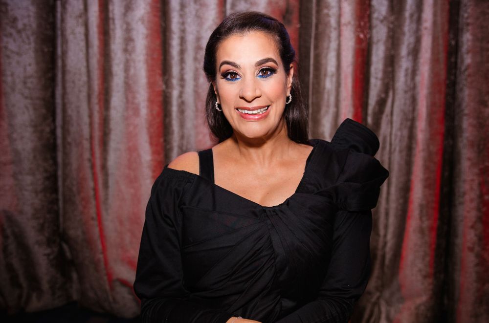 Woman wearing a black off-shoulder dress smiles while sitting against a dark curtained backdrop.