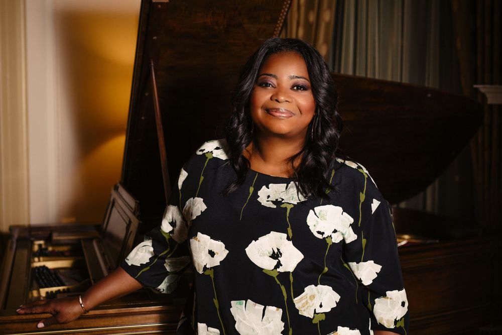 Octavia Spencer. Person in floral dress smiling warmly while seated by a piano.