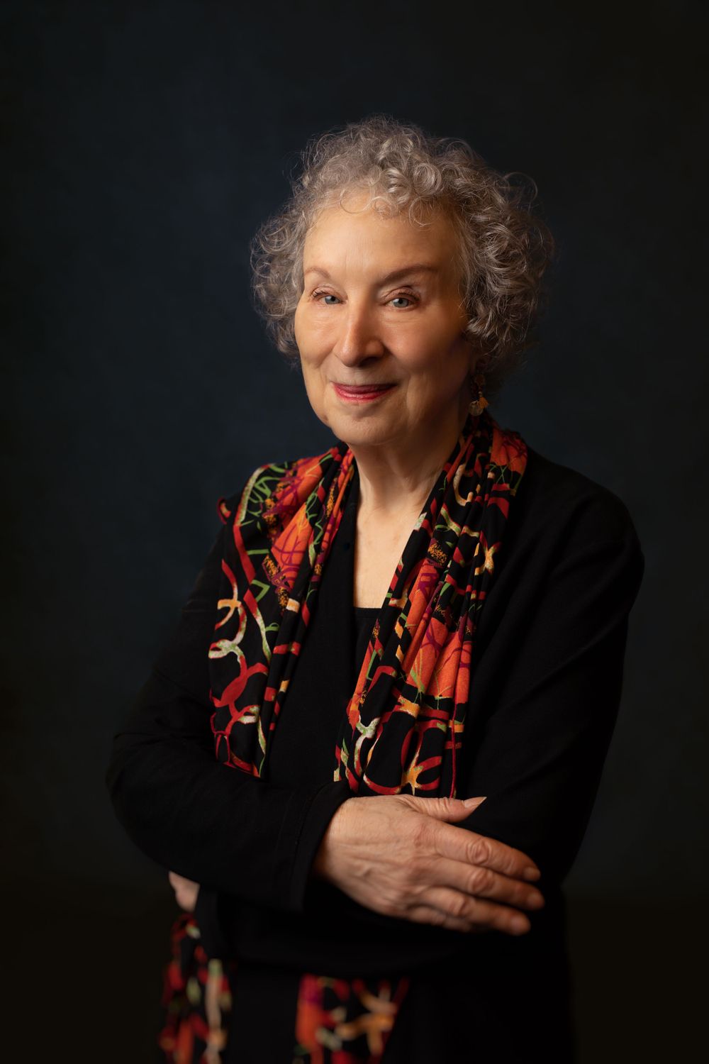 A woman with silver curly hair wearing a black top and patterned scarf poses against a dark background.