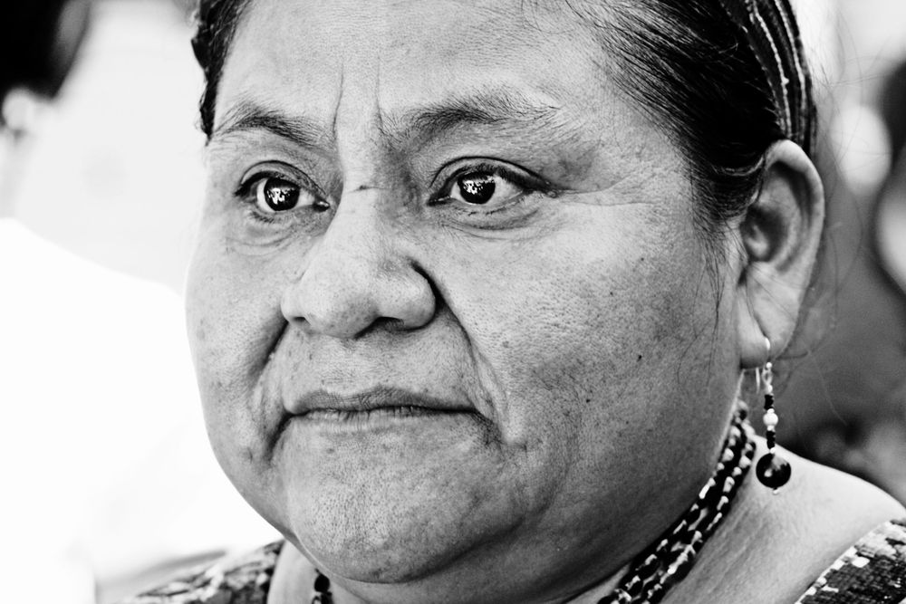A black and white close-up portrait of an Indigenous woman wearing traditional earrings and looking at the camera with a soft smile.