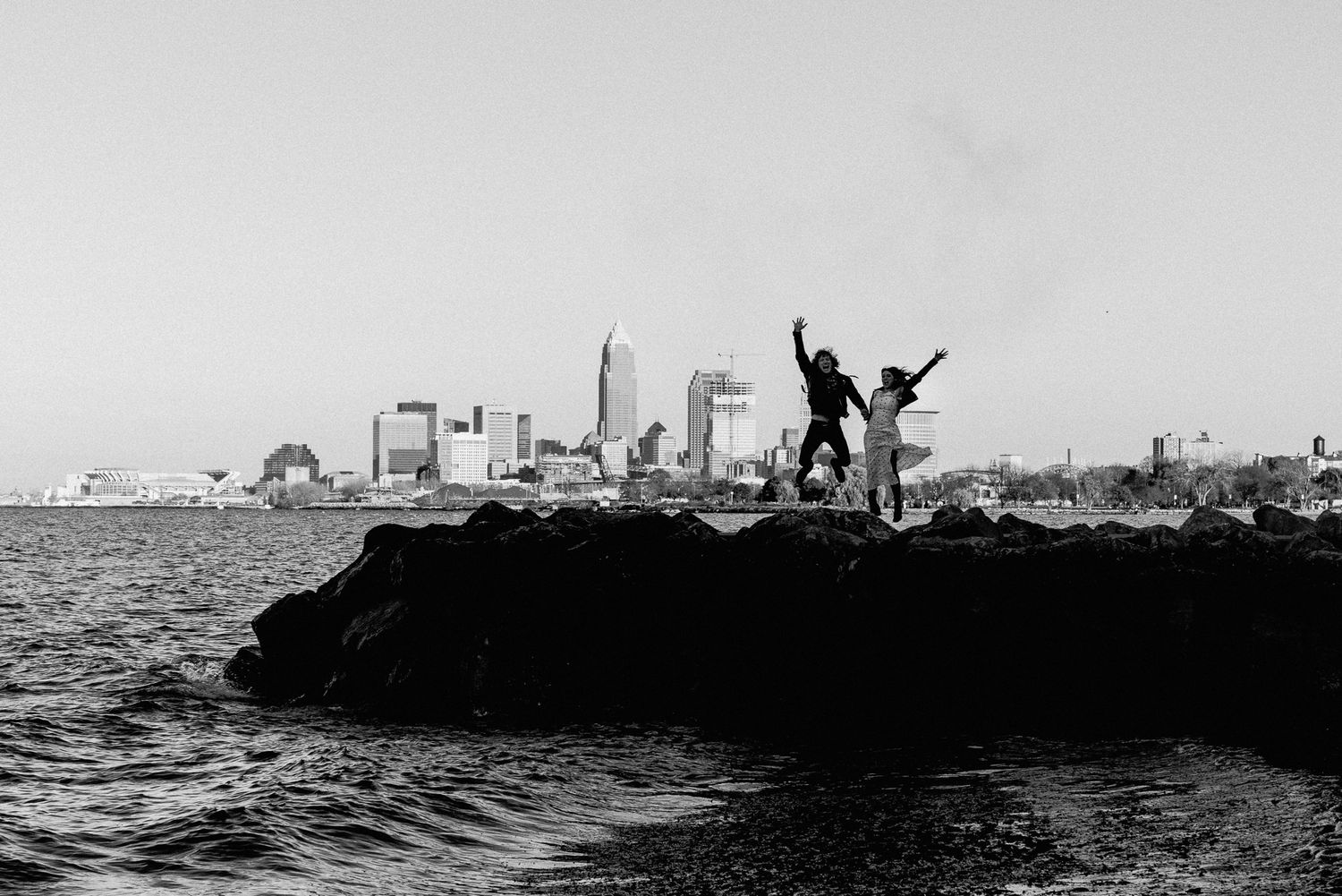 Engaged couple jumping on the rocks at Edgewater Beach with the Cleveland skyline behind them on their engagement photography session