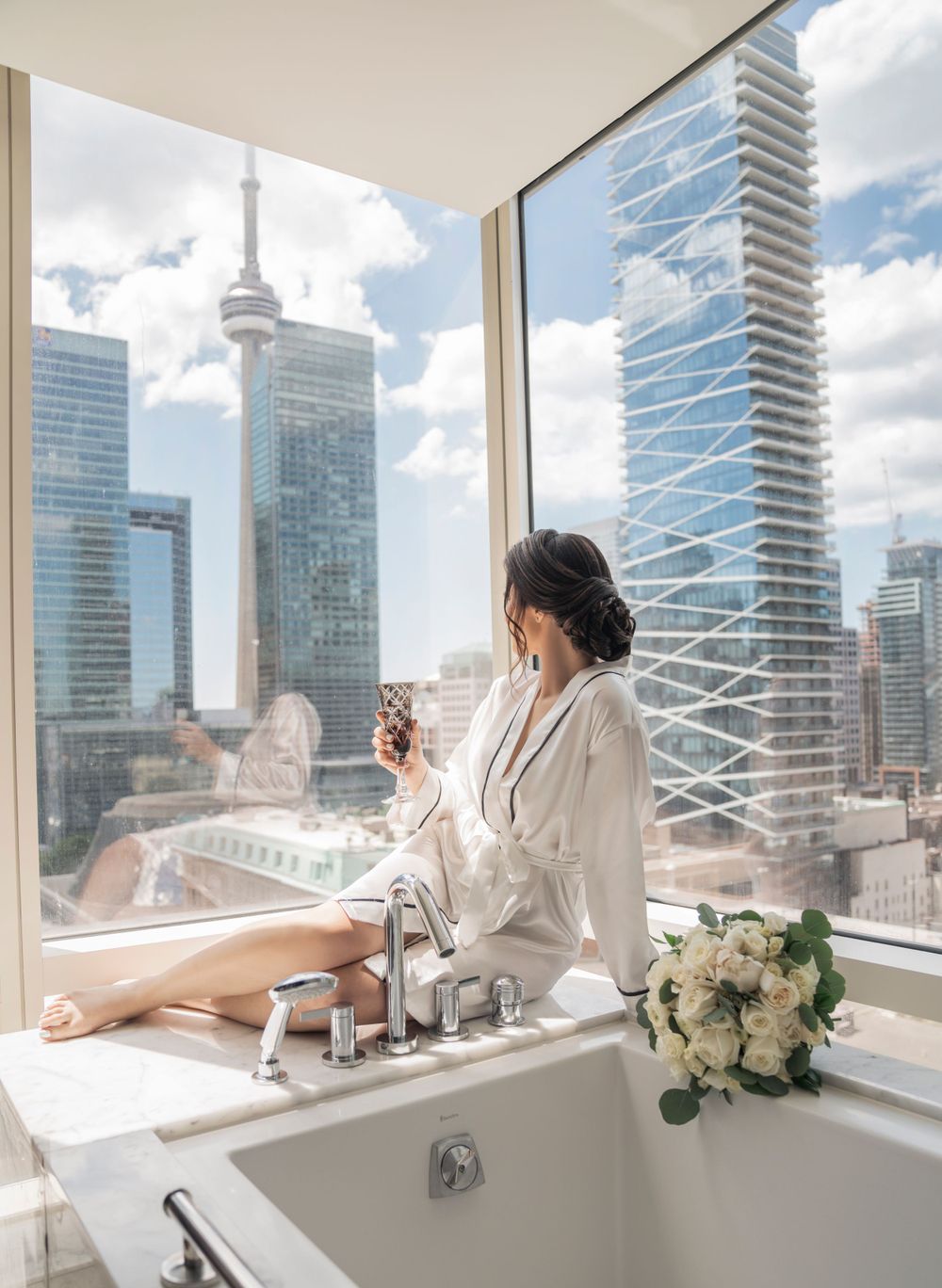 Woman in white robe enjoying morning coffee with Toronto skyline and CN Tower view from luxury hotel bathroom.