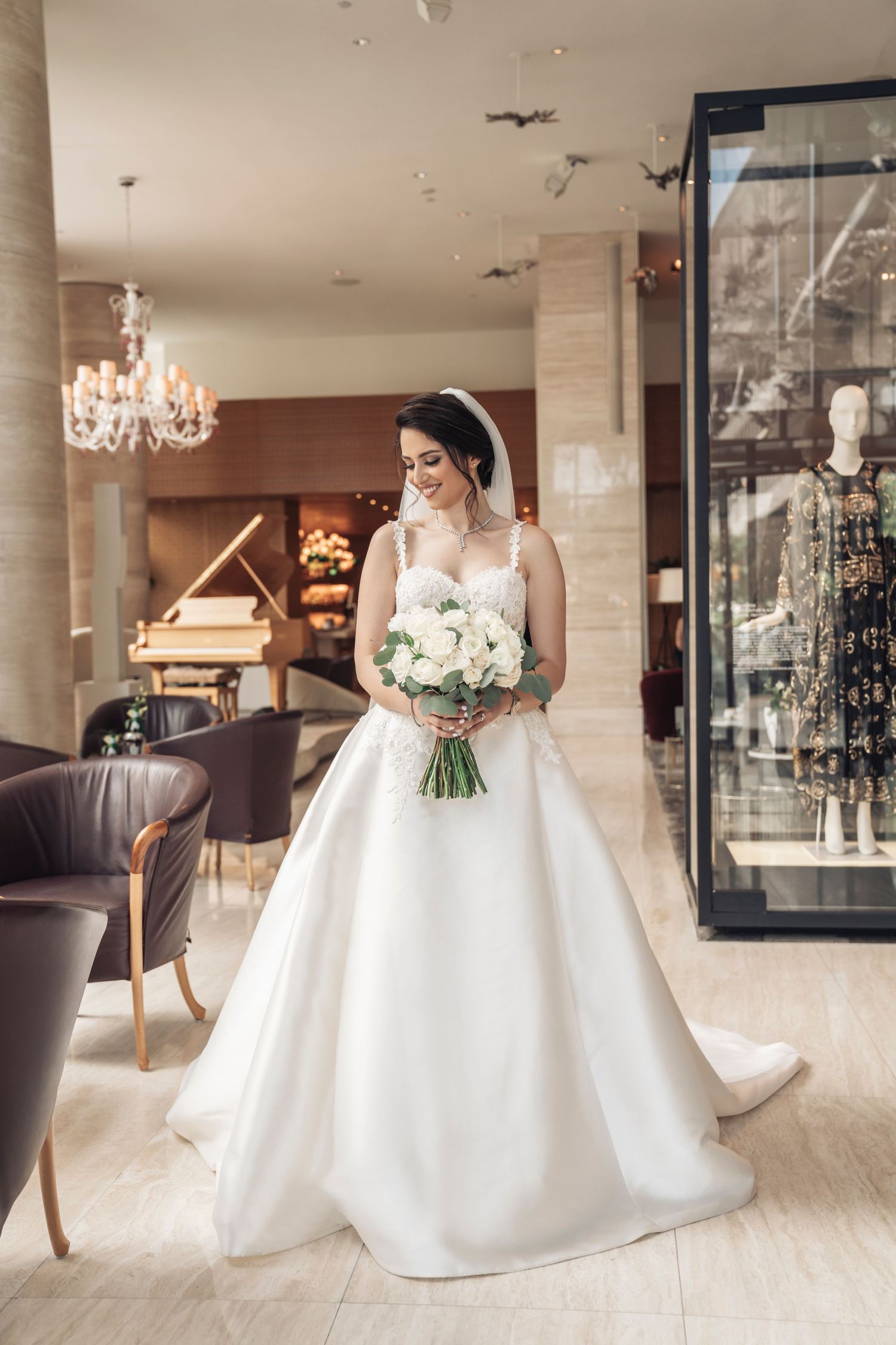 Bride in elegant white ballgown poses with bouquet in luxurious hotel lobby with chandelier.