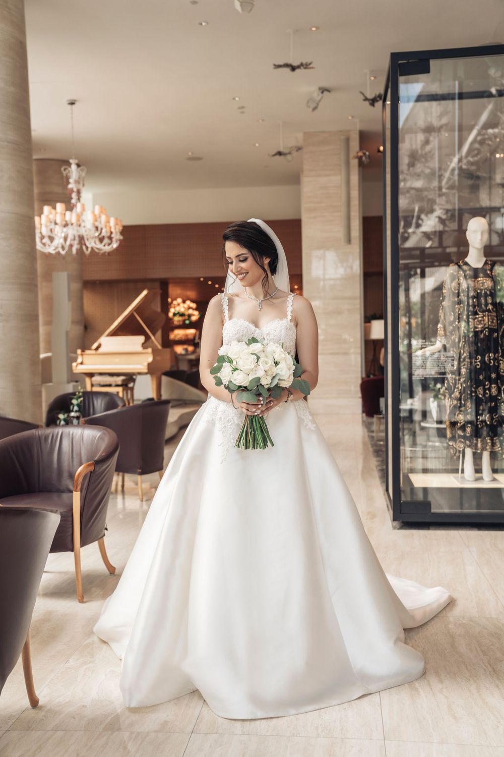 Bride in elegant white ballgown poses with bouquet in luxurious hotel lobby with chandelier.