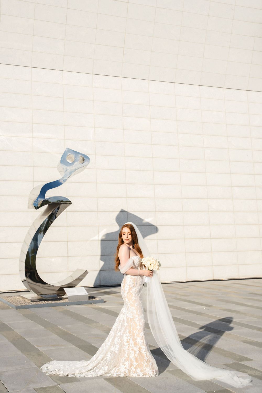 Bride in lace gown poses beside modern sculptural art installation against white building.