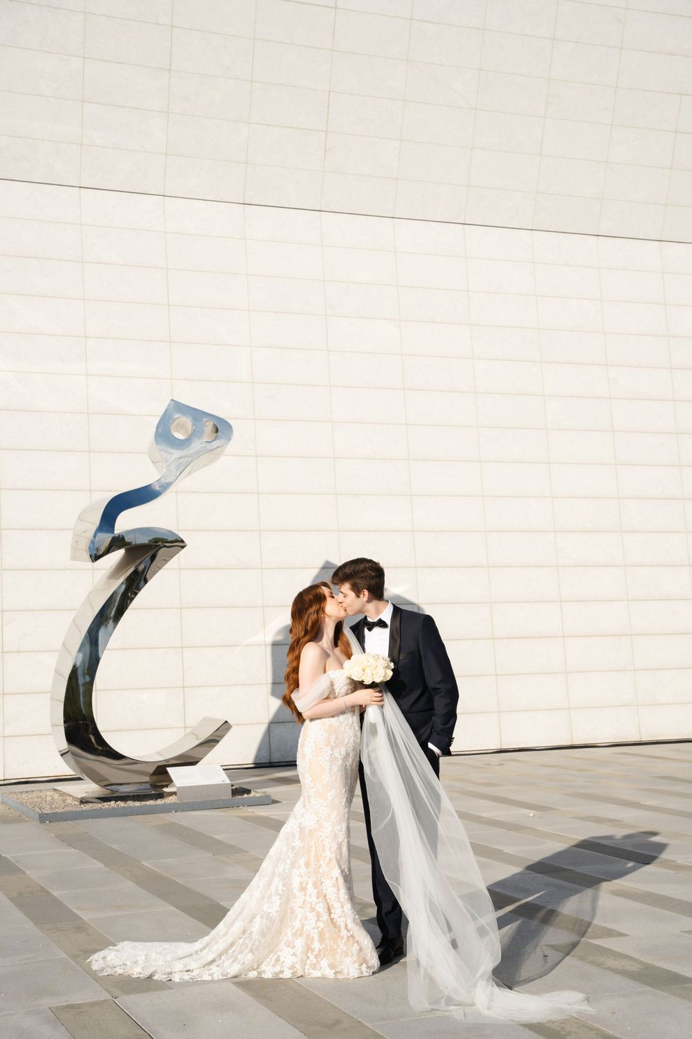 Intimate wedding moment shared between couple by modern sculpture against white architectural backdrop.
