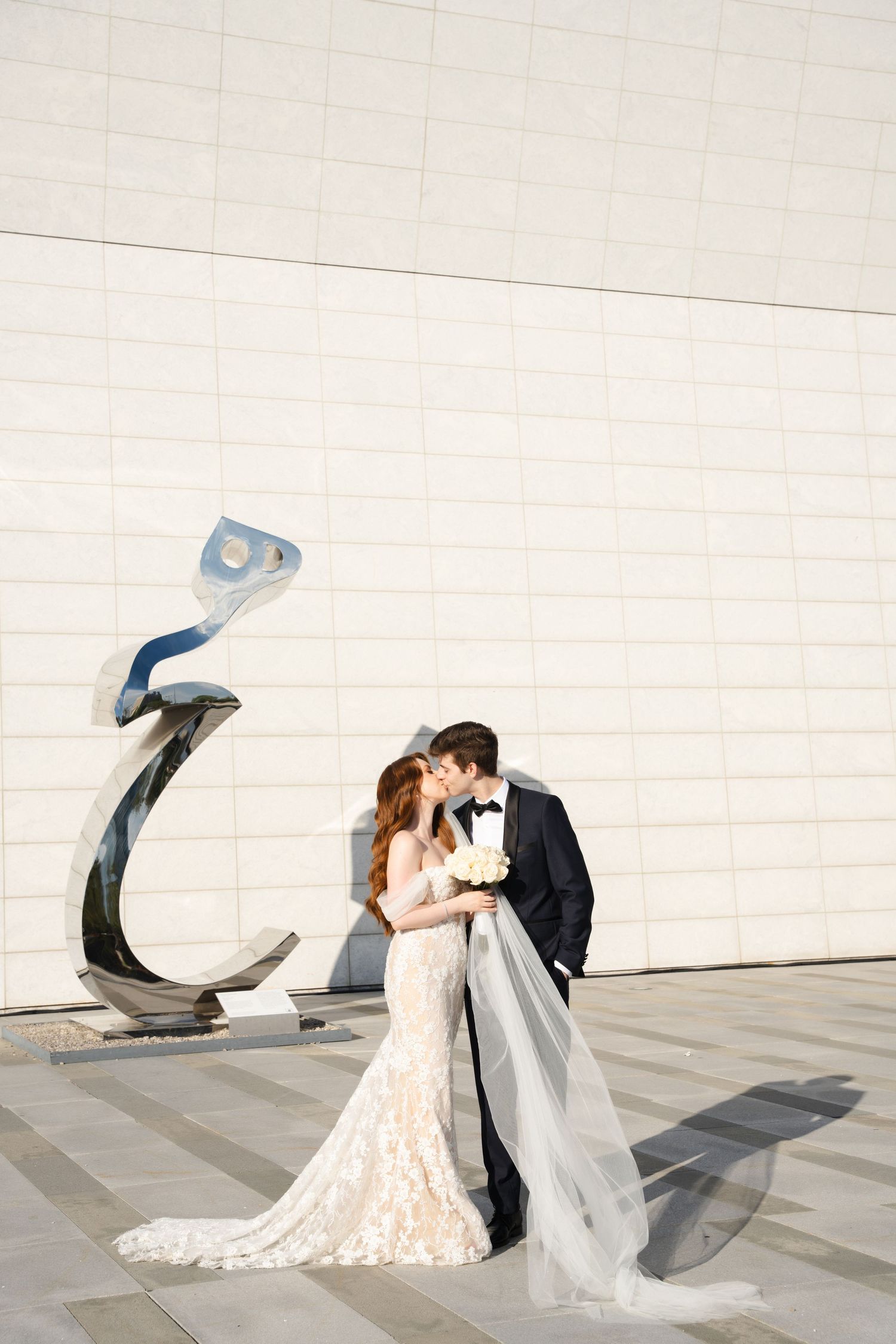 Intimate wedding moment shared between couple by modern sculpture against white architectural backdrop.