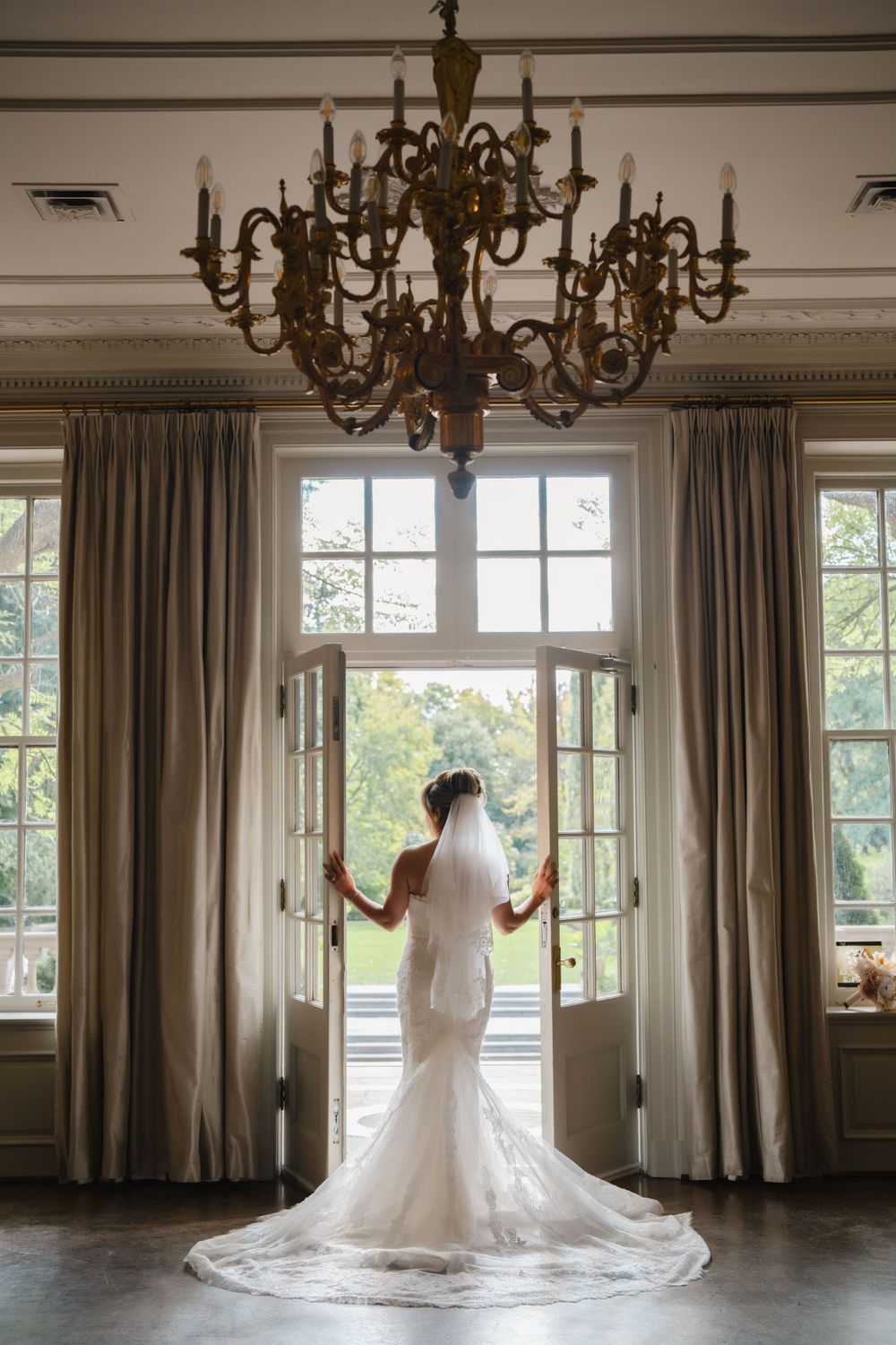 Bride in elegant white gown standing by window under vintage chandelier in grand room.