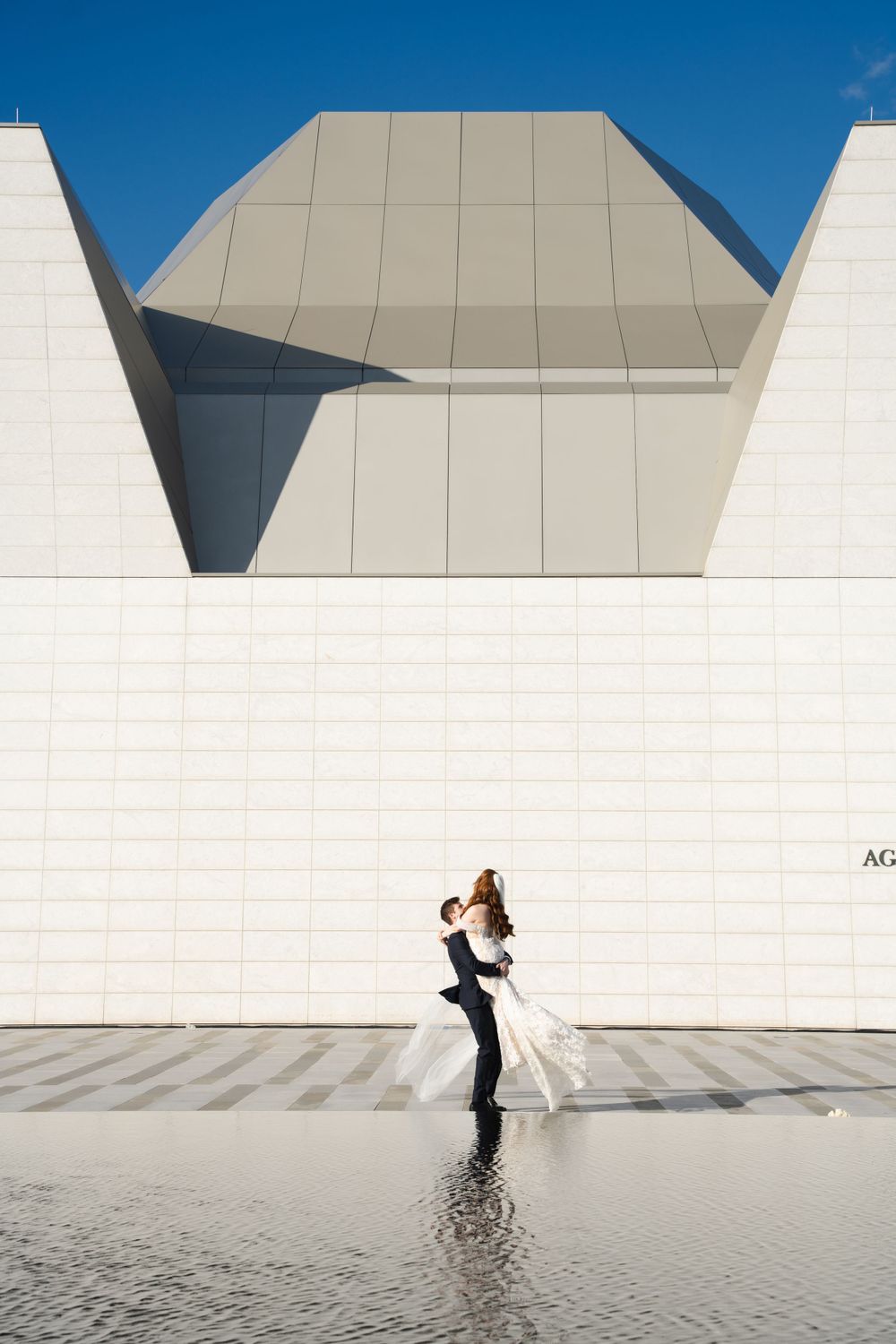 Wedding couple embracing in front of geometric white museum architecture with water reflection.
