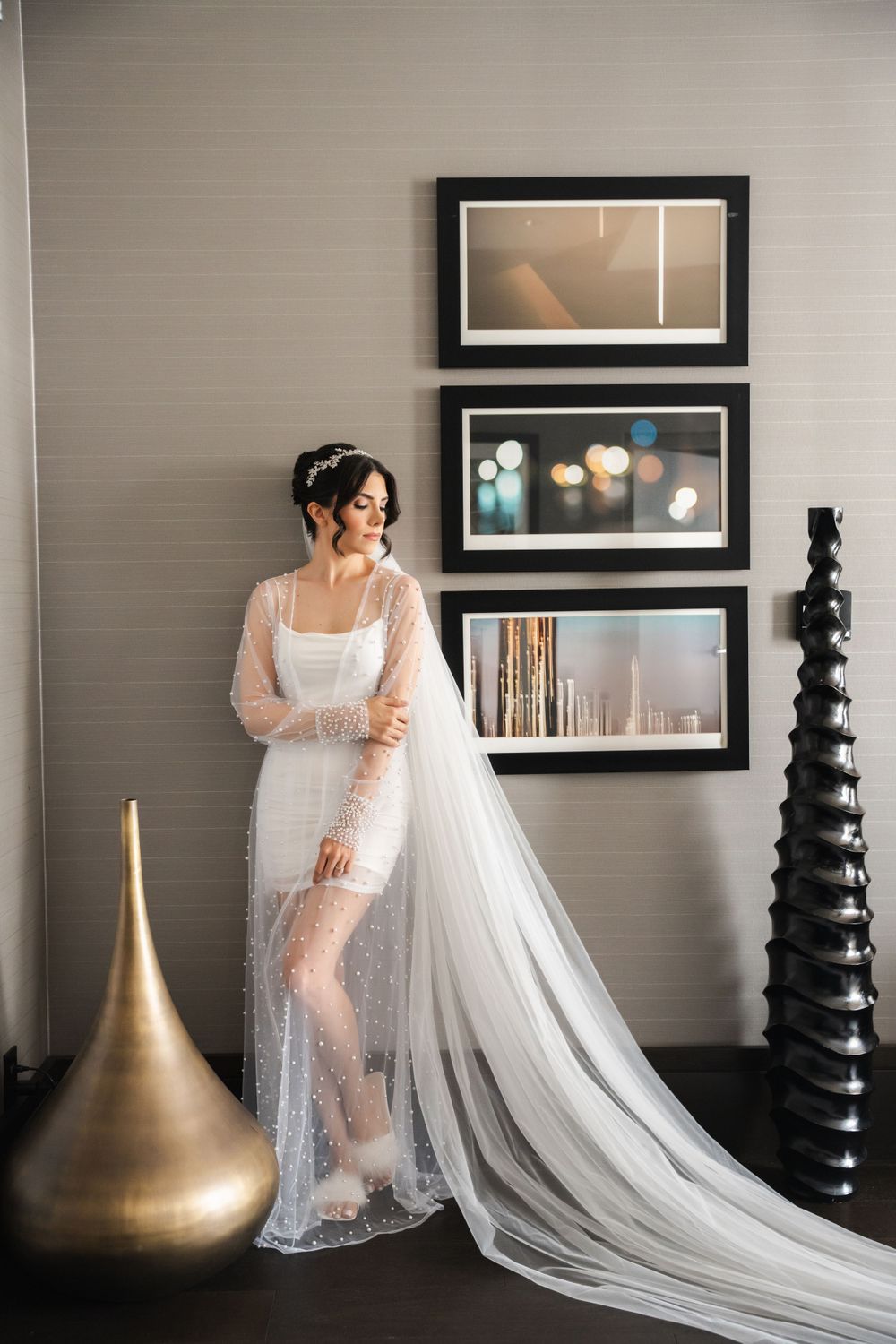 Bride in sheer lace gown poses against modern wall decor and gold accents.