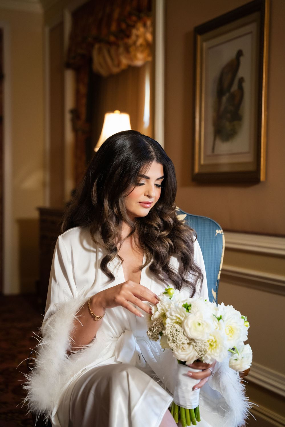 Bride in white feathered robe holds bouquet of white flowers in luxurious hotel setting.
