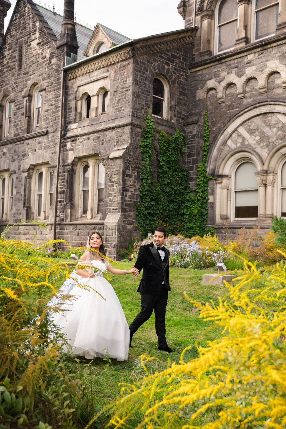 Couple poses in yellow wildflower garden outside Gothic stone building on their wedding day.
