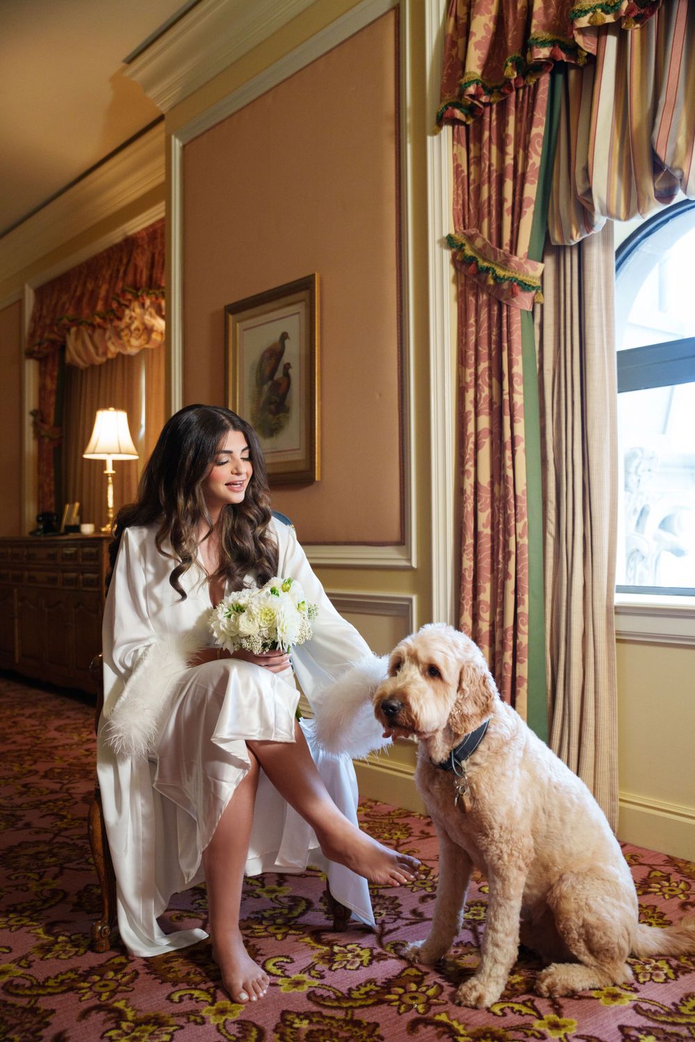 Sweet moment between a bride in white robe and her fluffy white dog in an elegant hotel room.
