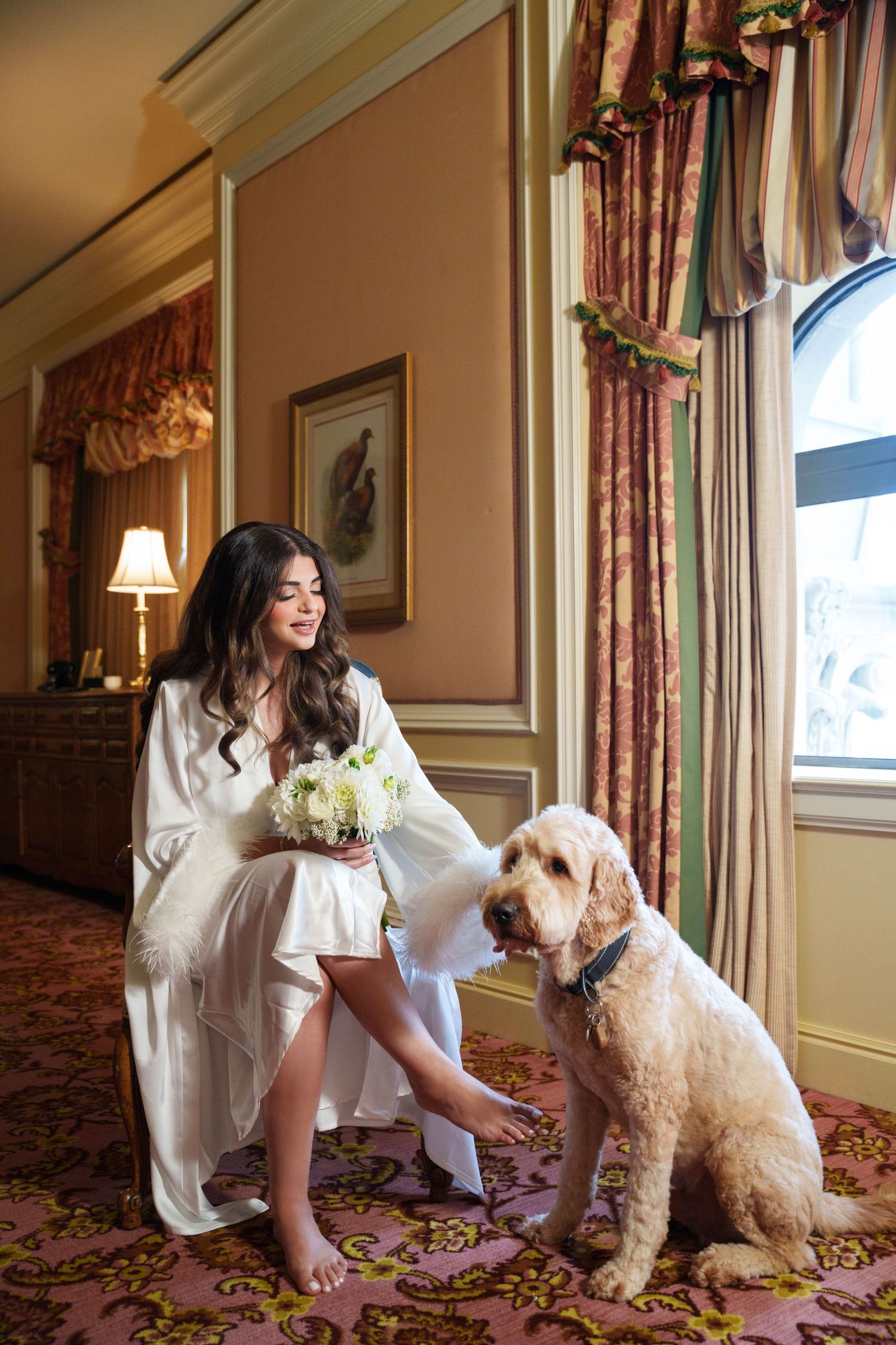 Sweet moment between a bride in white robe and her fluffy white dog in an elegant hotel room.