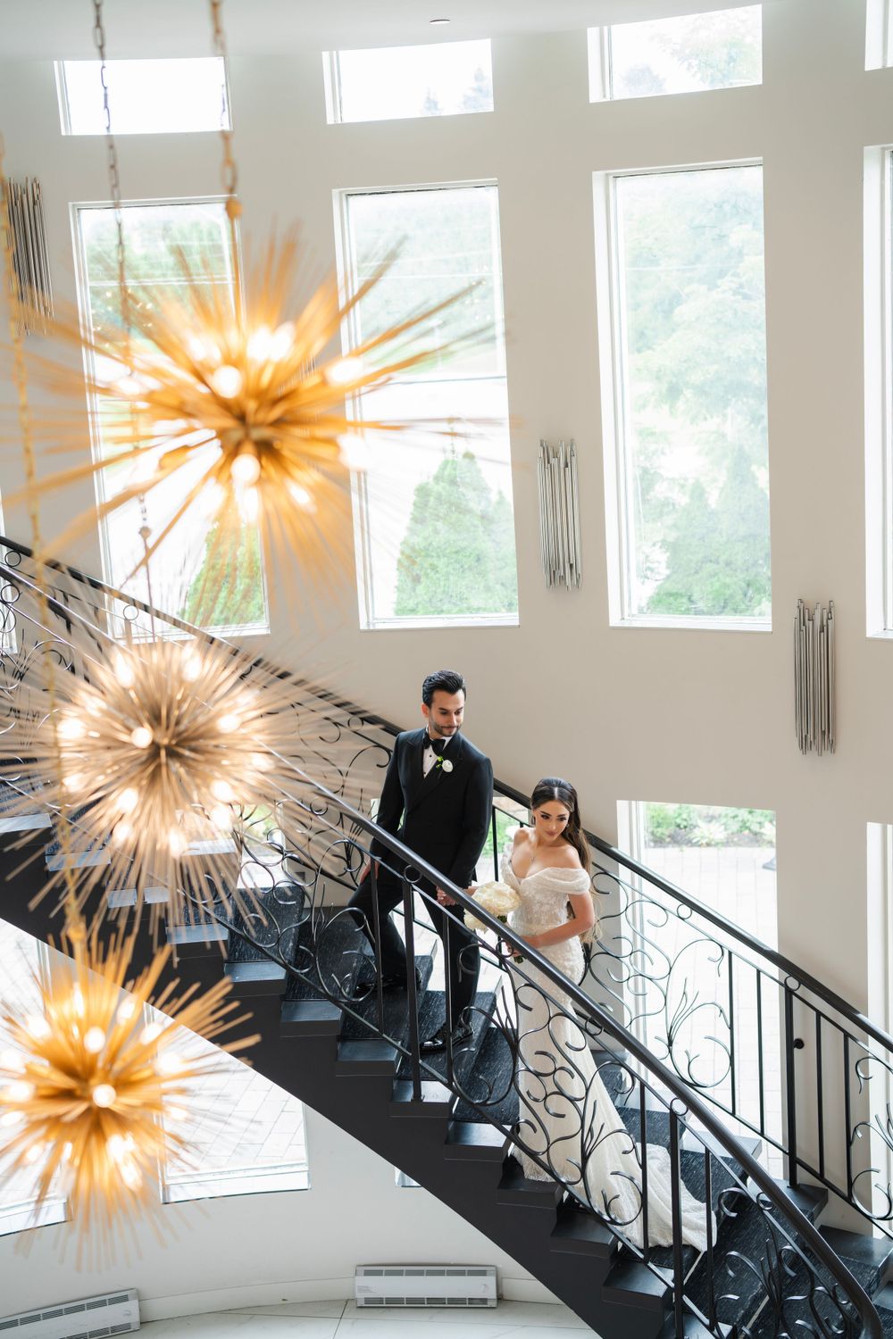 Elegant wedding couple descending grand staircase with dramatic starburst chandeliers overhead.