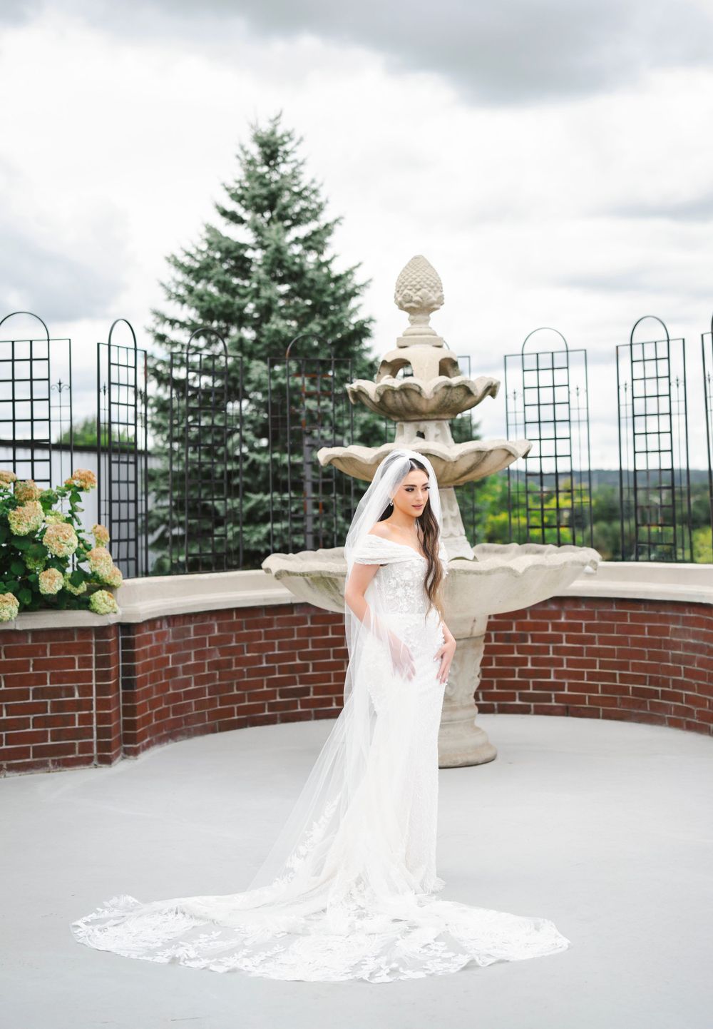 Bride in flowing white gown poses on terrace with brick wall and wrought iron fence.